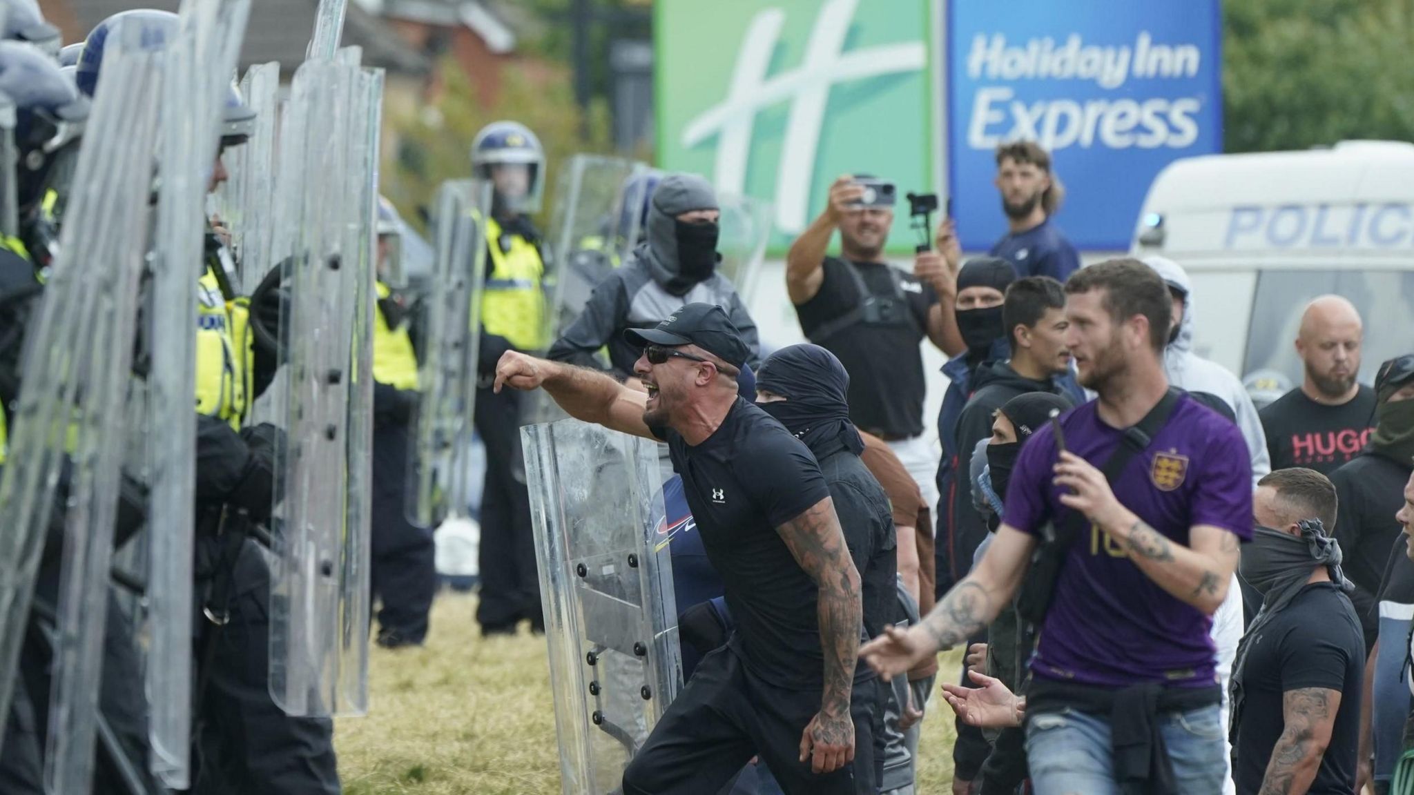 large crowd of men scream and shout at a line of police officers in riot gear and sheilds.