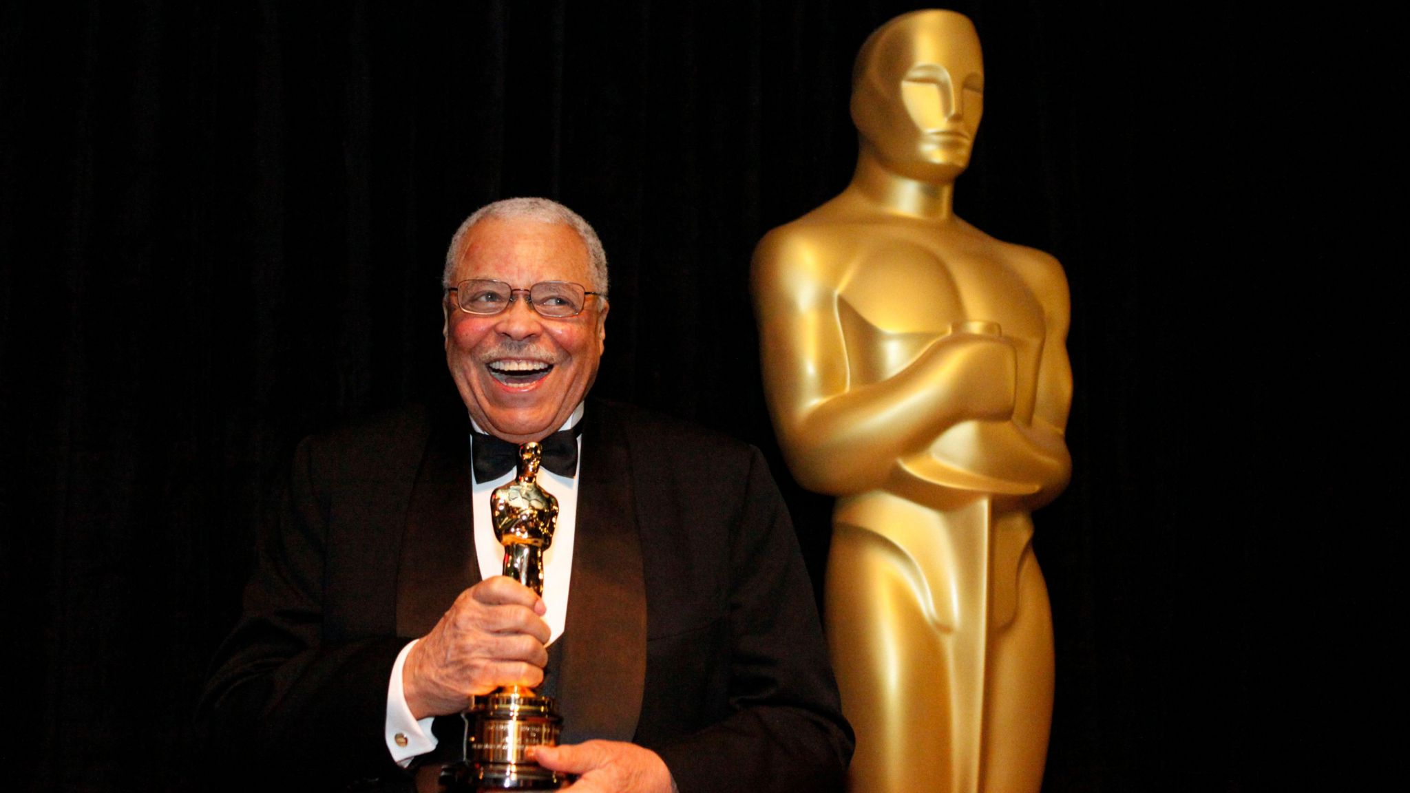 Wearing a black and white tuxedo with a black bow tie James Earl Jones smiles clutching his honorary Oscar next to a statue of an Oscar Award. This was taken at the 84th Annual Academy Awards show in Los Angeles in February 2012.