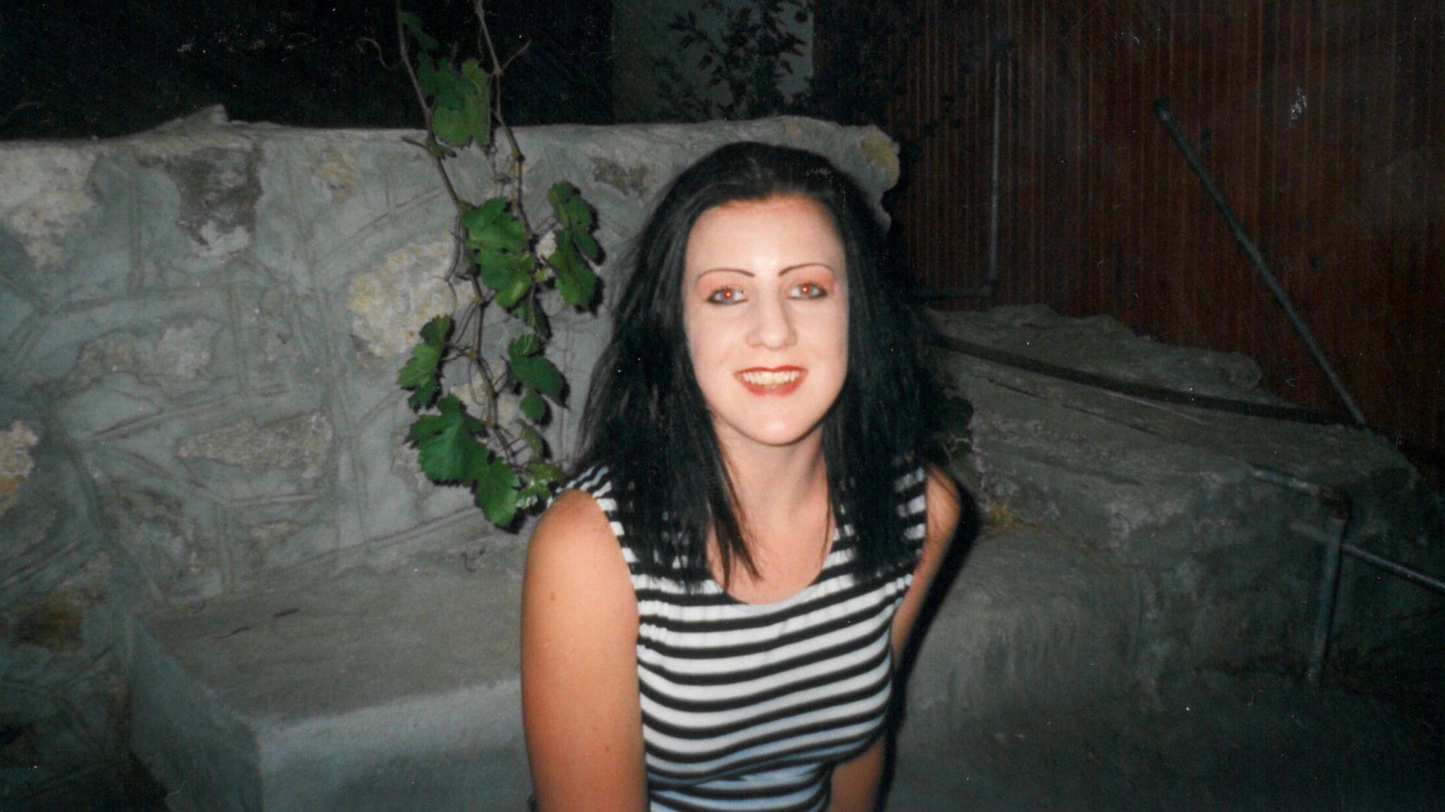 Anna, aged 17, smiles at the camera as she sits on a step. She has long dark hair and is wearing a black and white striped vest.