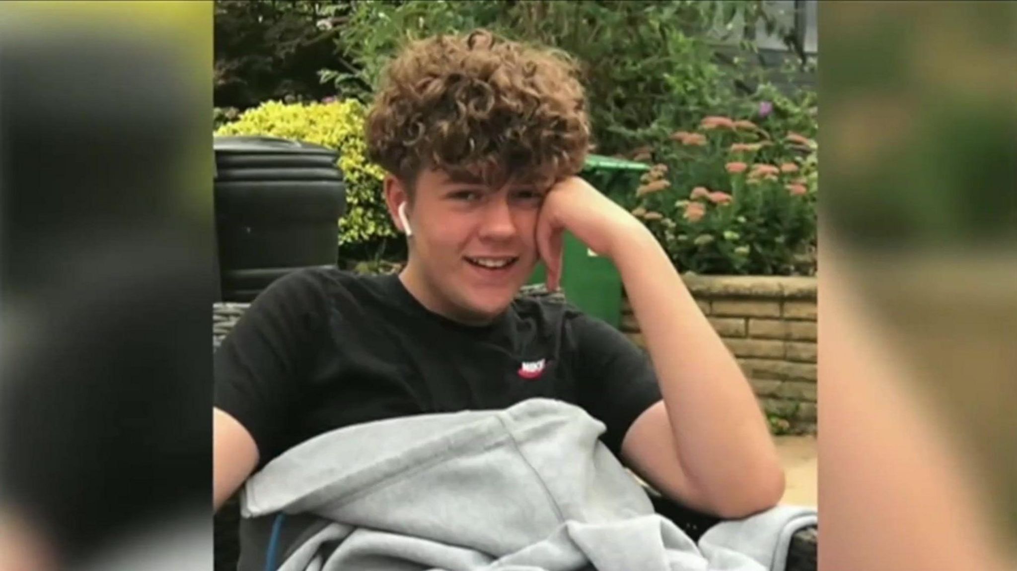 A boy with curly hair wearing a black t-shire, sat down in front of shrubbery