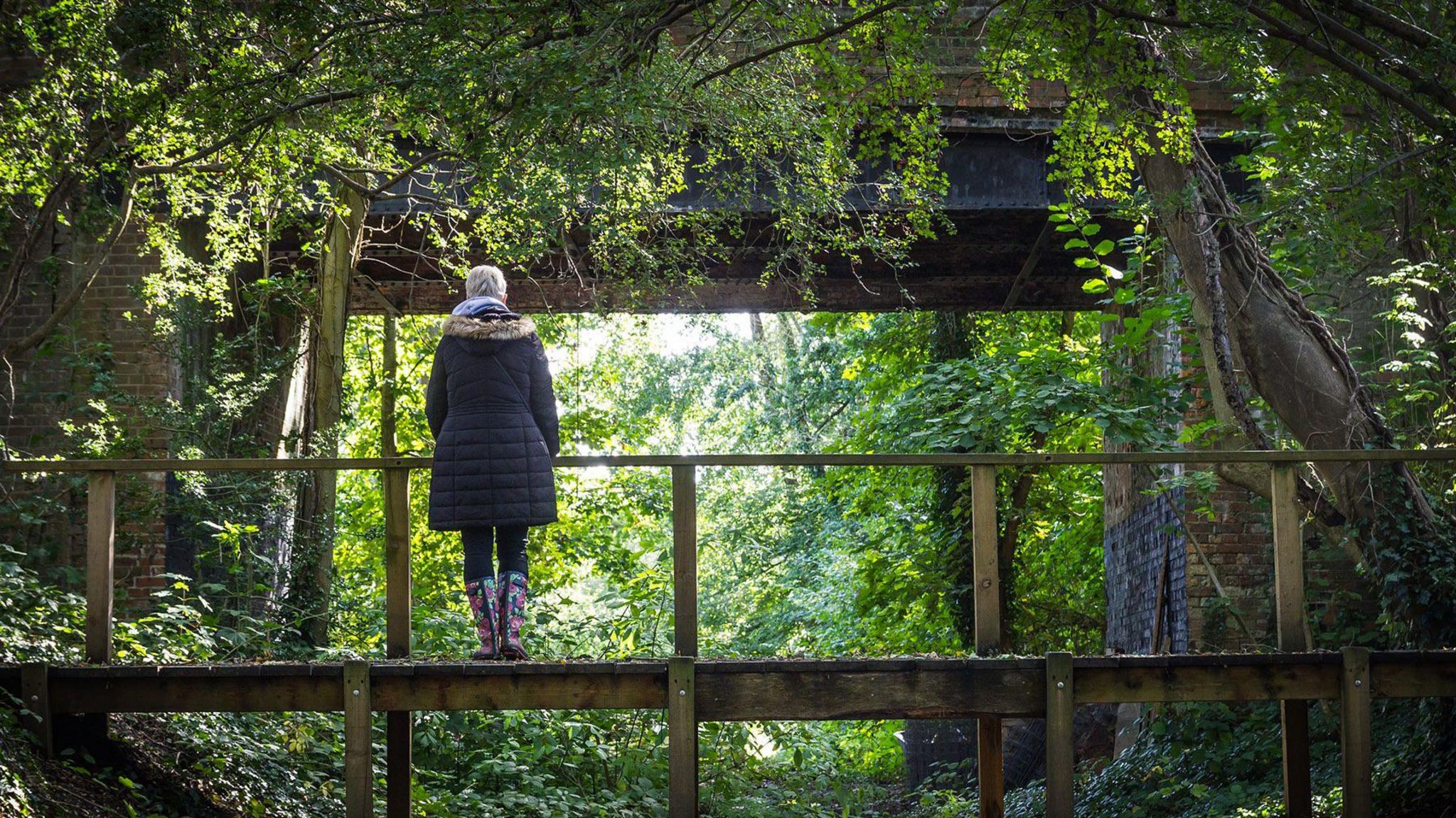 A woman with short white hair and a long black coat stands on a small wooden bridge looking away from camera at the Barcombe bridge structure which is surrounded by trees