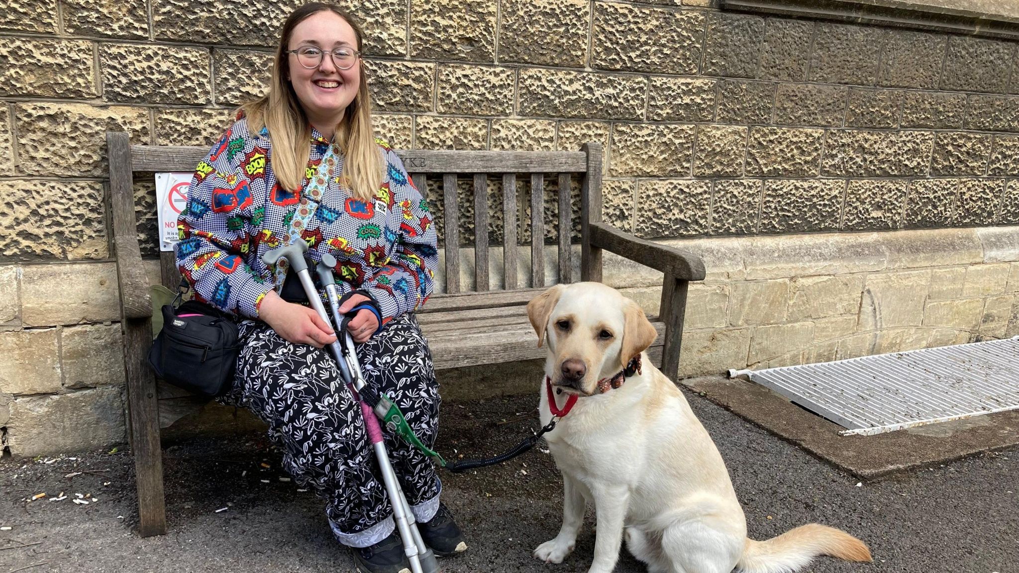 Martha Hall sat on a bench wearing a colourful outfit. Her yellow Labrador, Greg, is sat next to her on the floor.