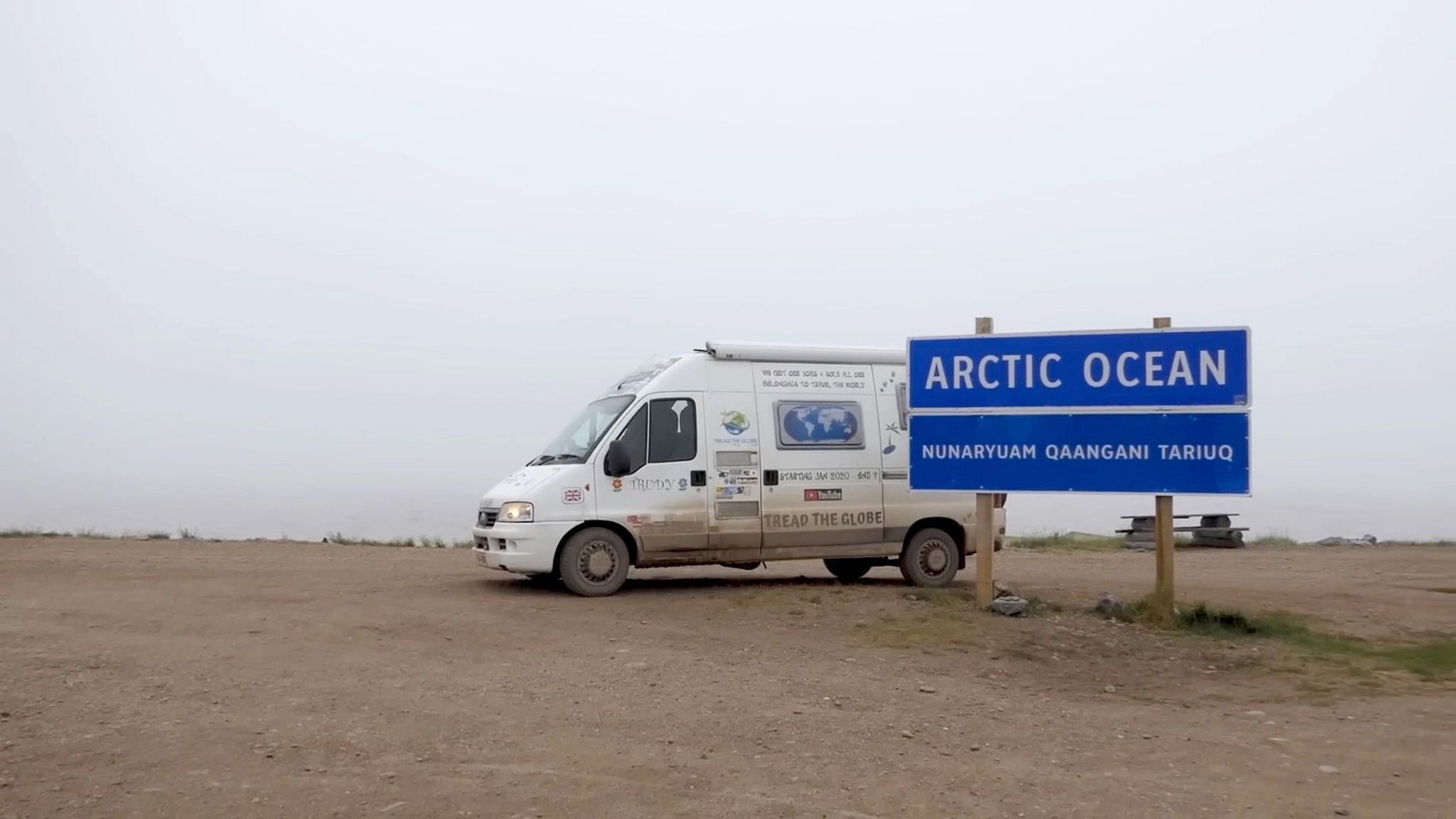 A white camper van parked on a barren dirt patch in front of a blue sign that says Arctic Ocean