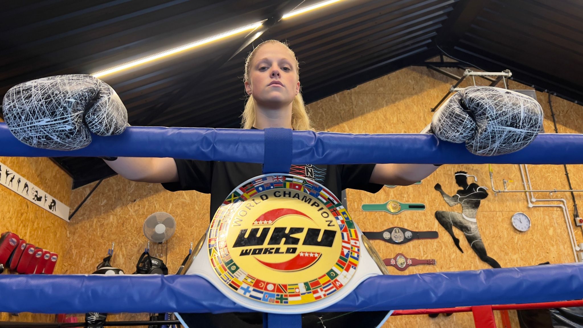 Hannah Turner is pictured staring down at the camera with a powerful facial expression. She is standing at the edge of a boxing ring with her boxing gloves on, resting over the ring's padded edge. Her world champion belt rests in front of her.