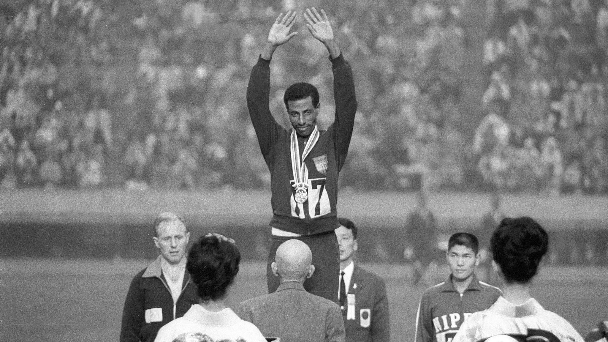 Abebe Bikila standing with his arms raised on a podium after winning his second Olympic gold