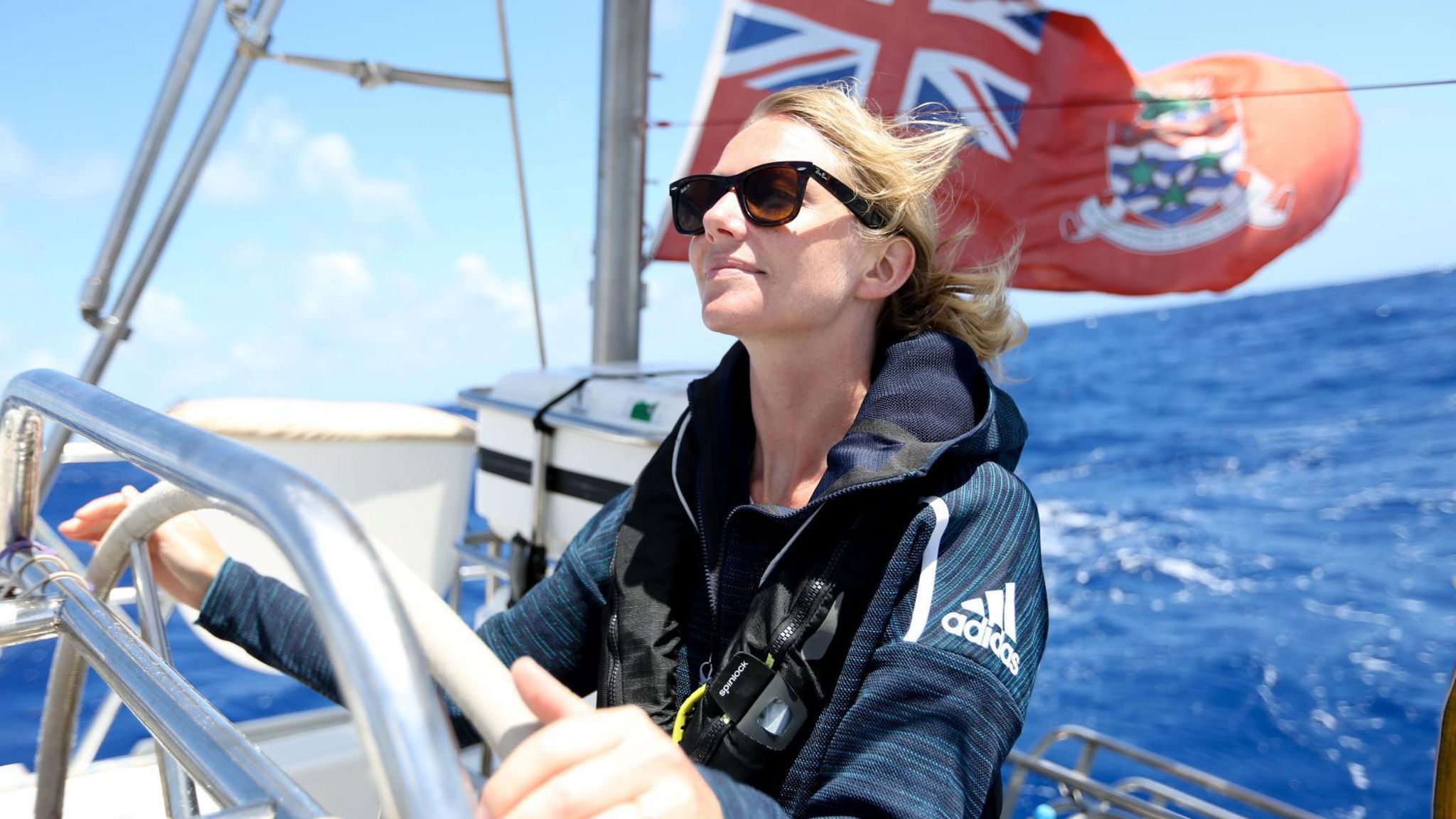 Emily Penn. A woman sailing a boat in the North Pacific. She is blonde and wearing sunglasses