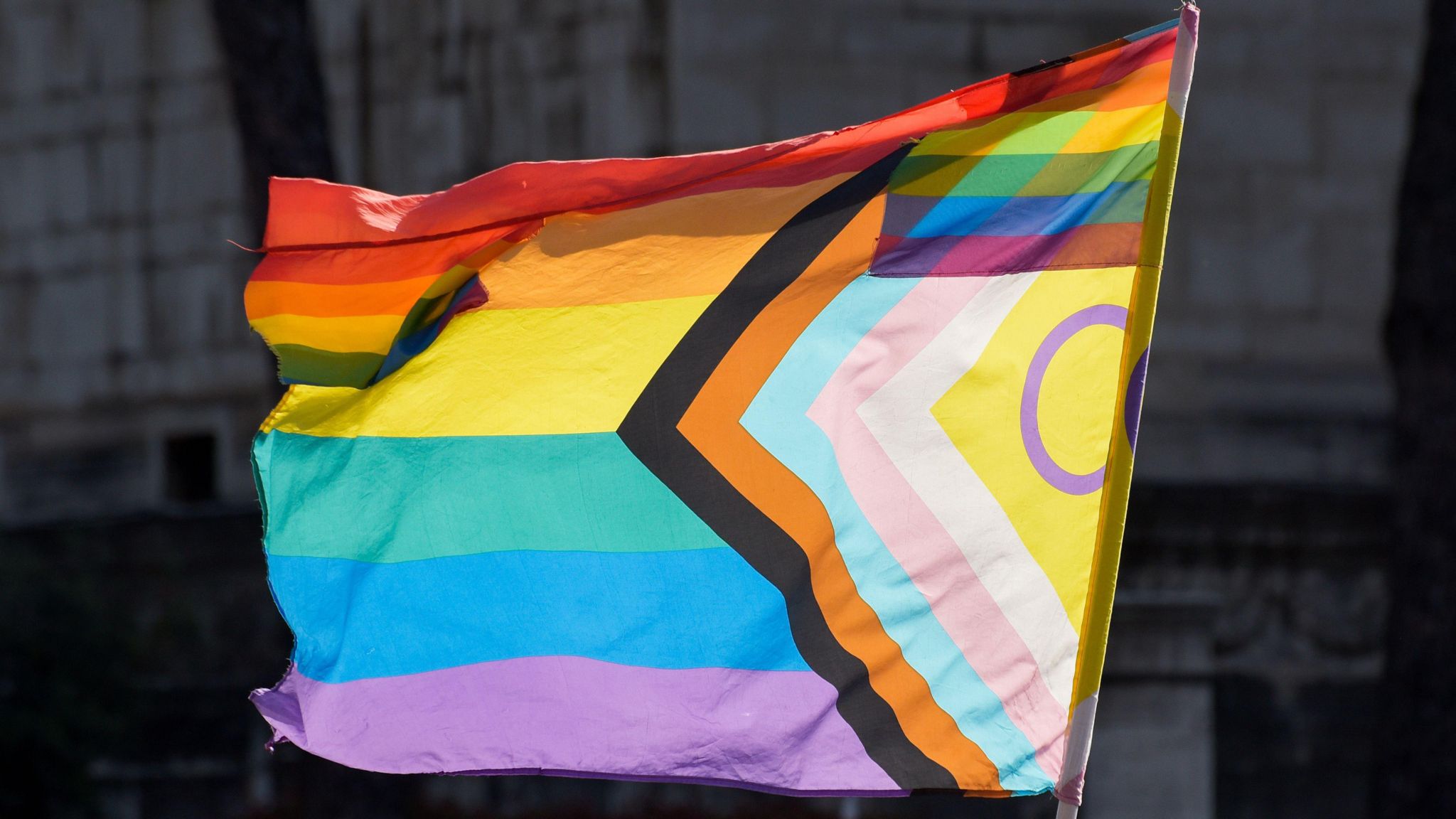 Image of the progress pride flag. It has rainbow stripes and a triangle on the side.