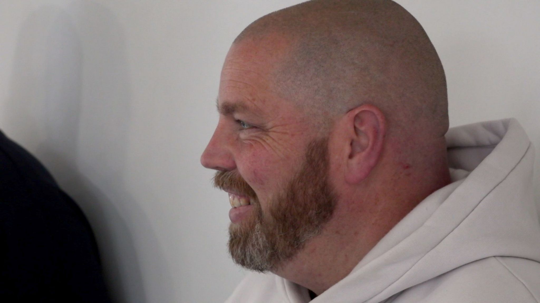 Paul Barran, a white middle aged man with his hair shaved and a ginger beard wears a white hooded jumper and smiles looking away from the camera