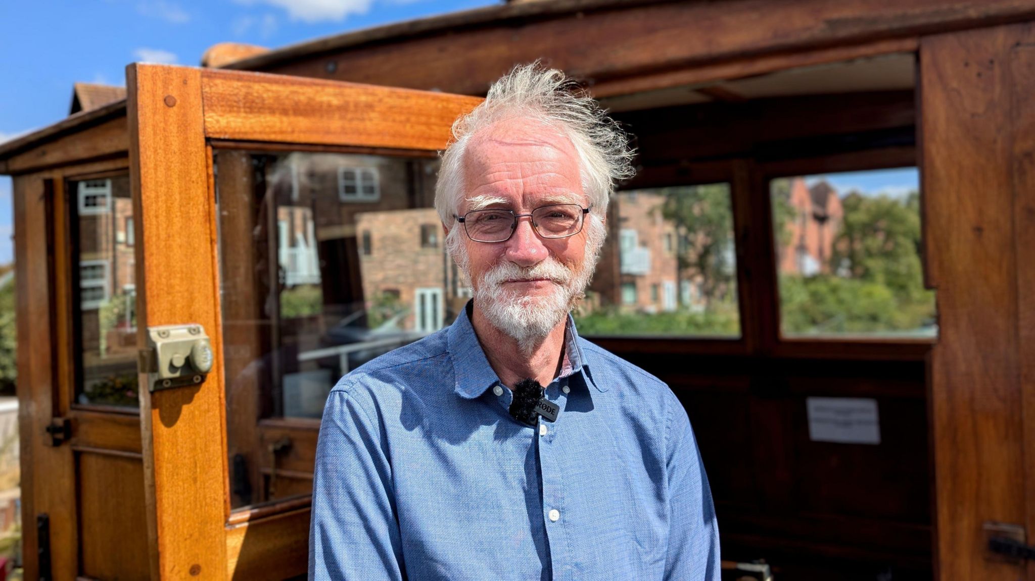 Iain Campbell smiles into camera while standing in the doorway of Sytan's wheelhouse. He is wearing a blue shirt and there are terraced houses and a bright blue sky visible behind the small structure.