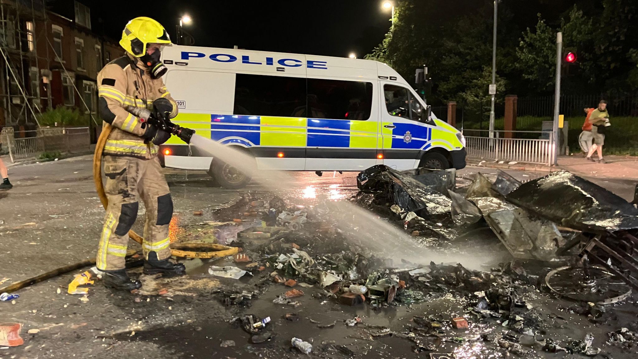 A firefighter extinguishes flames from the rubble of a bonfire, following the riots in Harehills, Leeds. A police van stands across the road in the background