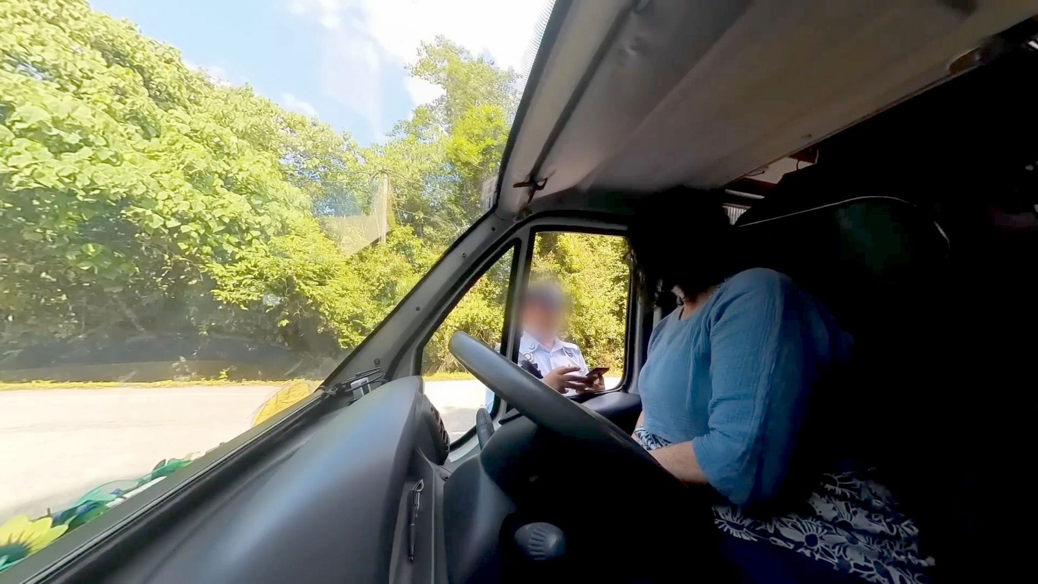 A view inside a camper van of a woman in a blue top talking to someone through the driver's side window, their face has been blurred out. Behind them trees are visible