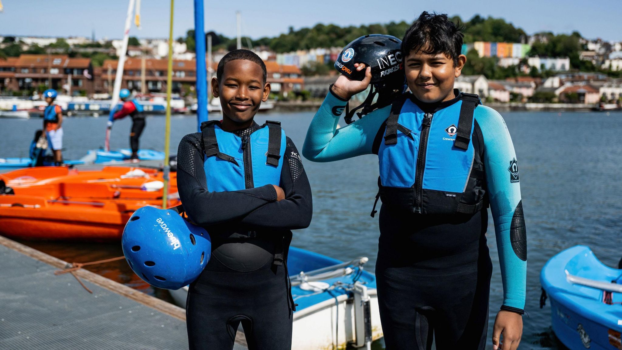 Two young boys standing next to the water in front of a few small boats. They are in blue life jackets and are holding blue helmets and smiling