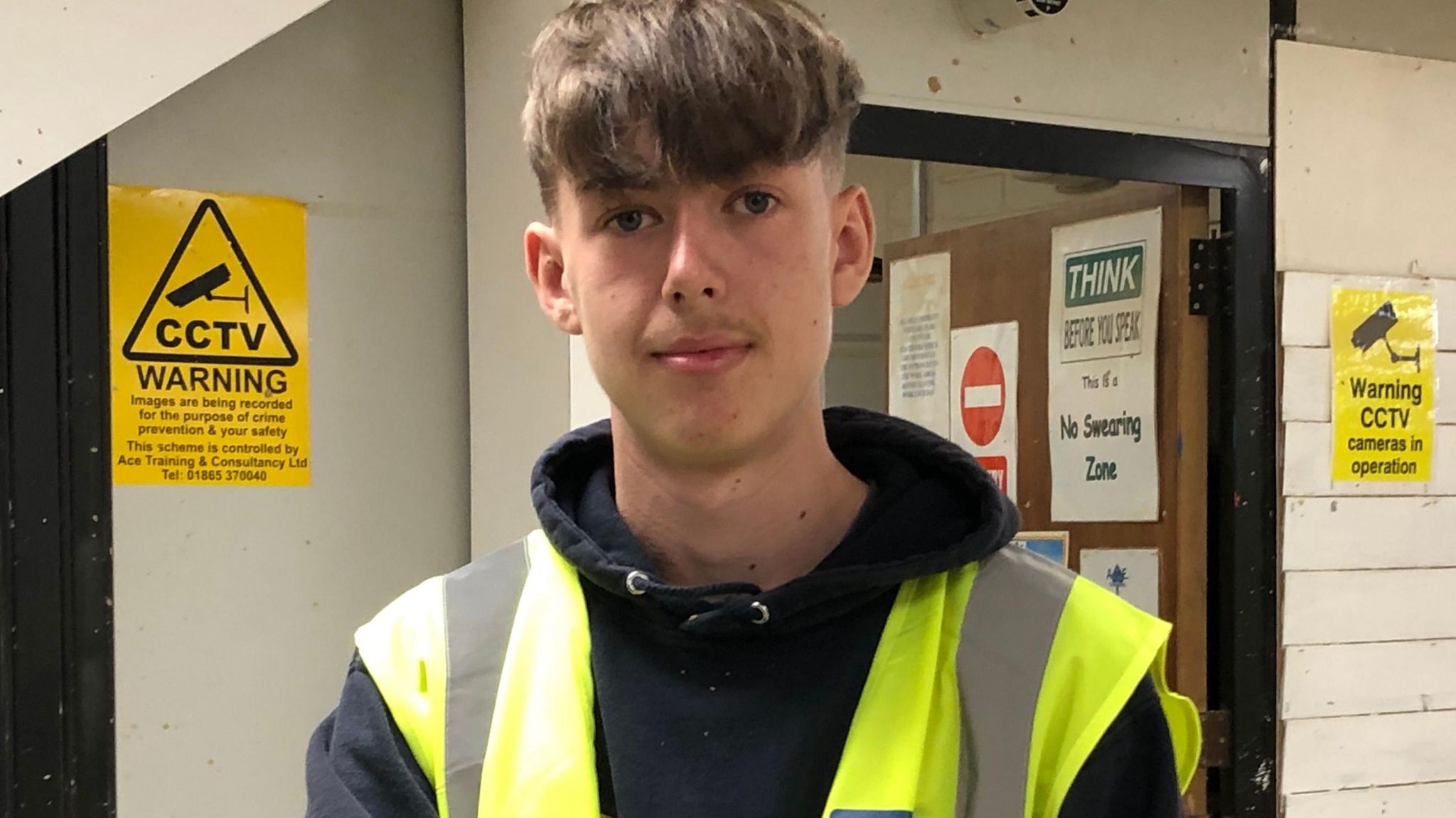 Riley has brown hair with a long fringe and is wearing a black hoodie with a high-vis sleeveless jacket over the top. He is standing inside a workshop in front of an open door with yellow posters either side warning CCTV is in operation. Another sign says it is a no swearing zone