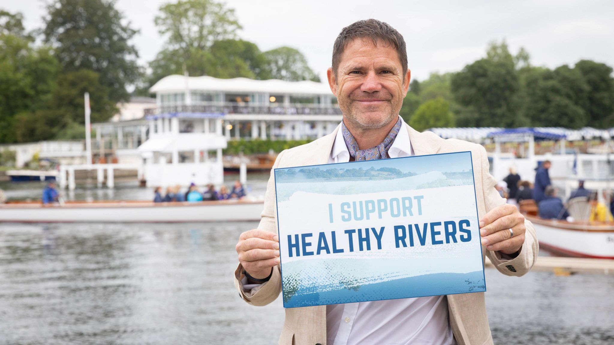 A dark-haired man in a cream blazer and white shirt stands in front of a river holding up a sign saying 'I support healthy rivers'