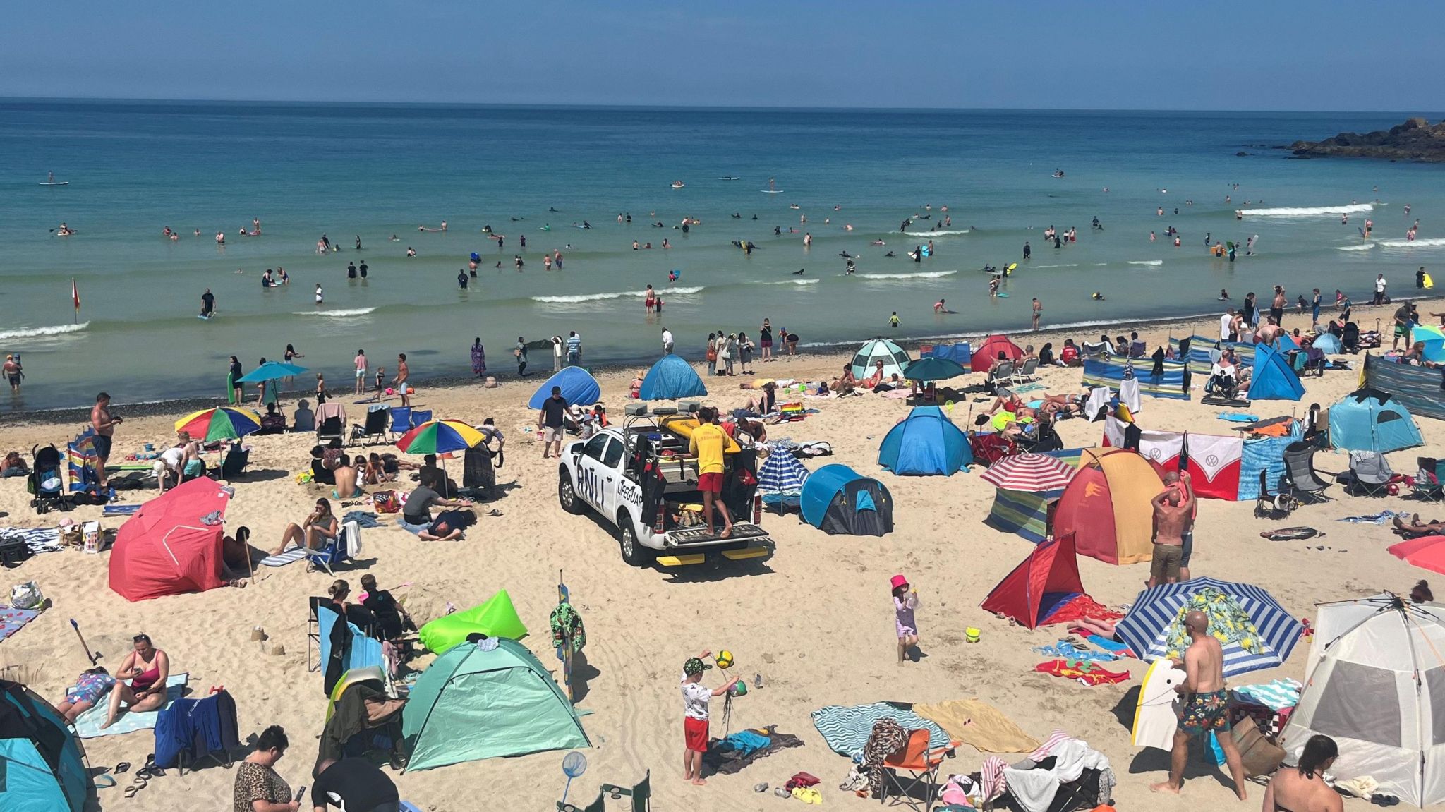 A lifeguard pickup truck in the middle of a busy Porthmeor beach which is corwded with people and colourful tents and umbrellas, the bule ocean with small waves is visible in the background