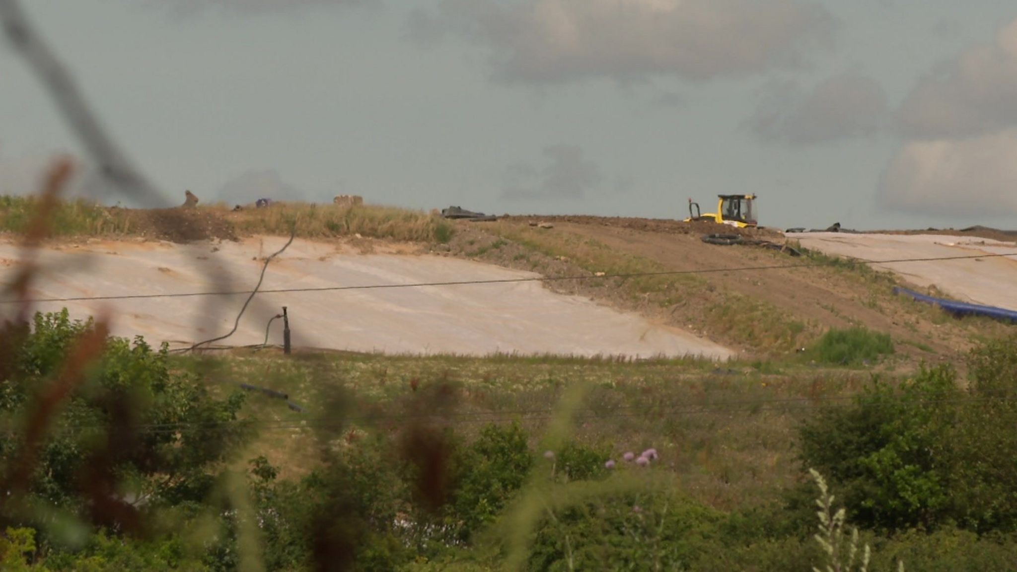 A yellow construction vehicle is seen in the distance atop the hilly landfill site.