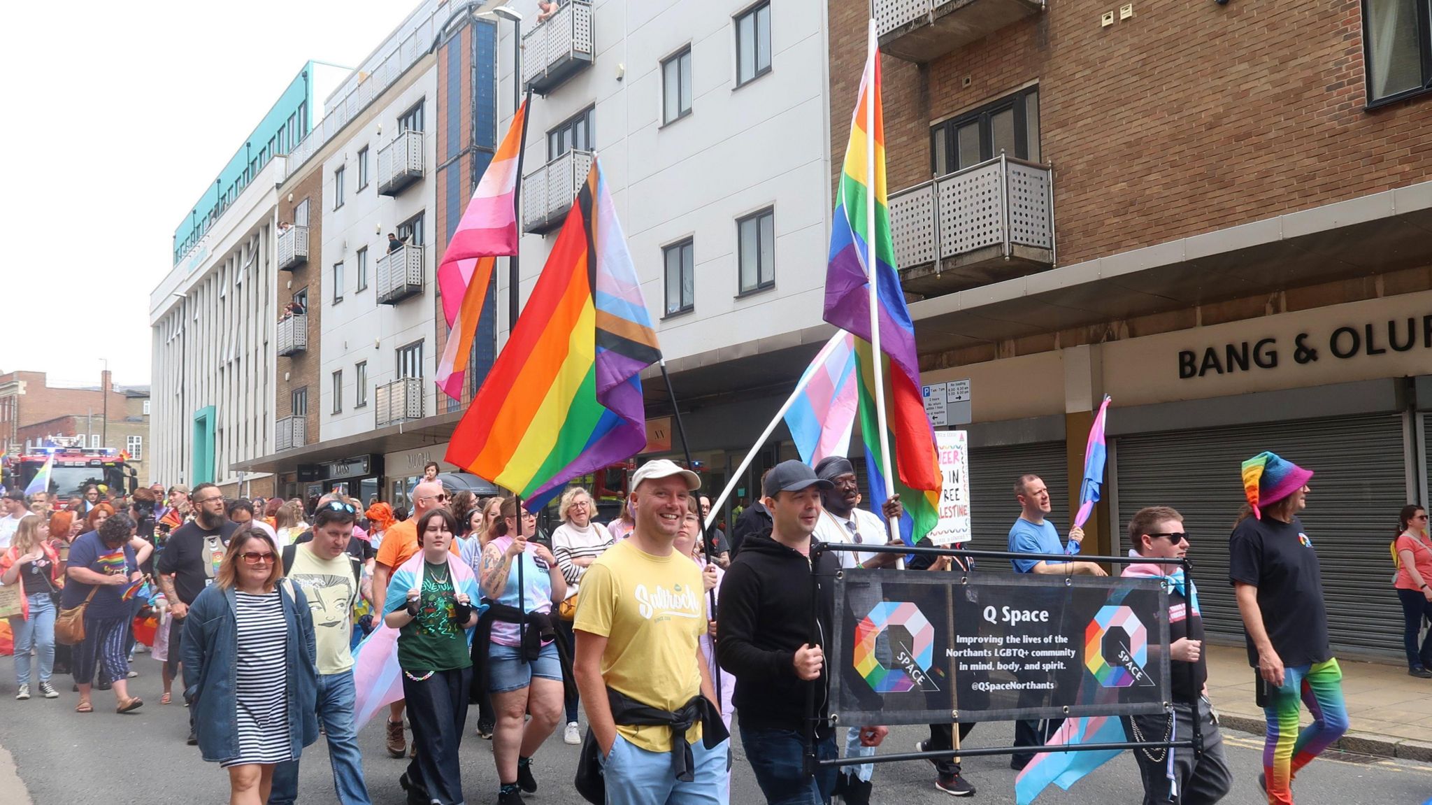 A large group of people walking through the streets of Northampton holding rainbow flags