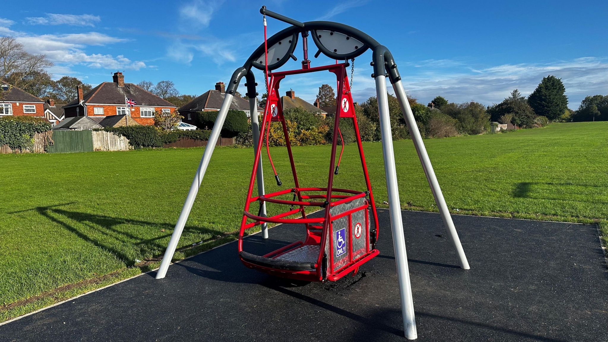 A red children's disability swing, it is on a tarmac base with the grassy park in the background