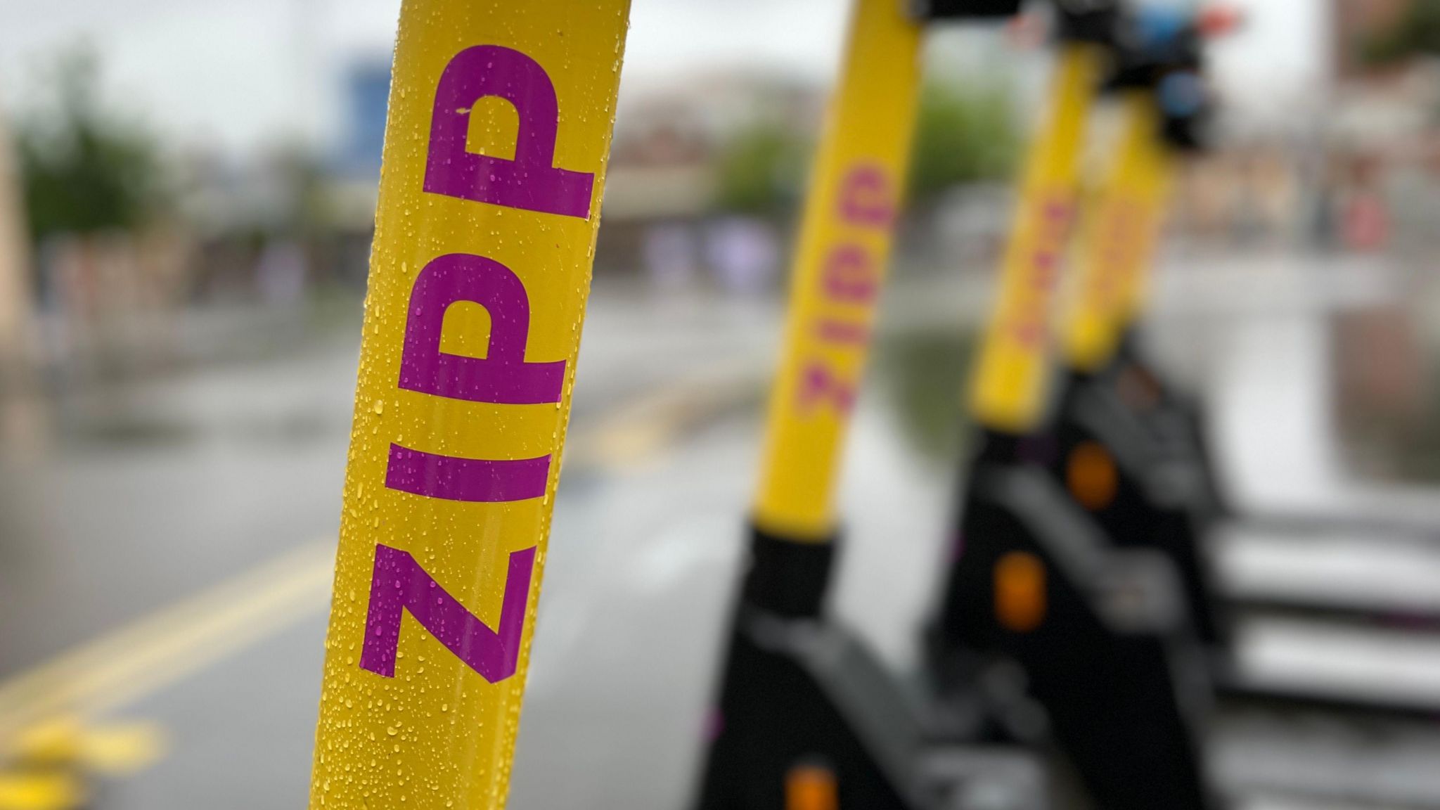 A Zipp logo in pink on the yellow body, with other e-scooters in the background