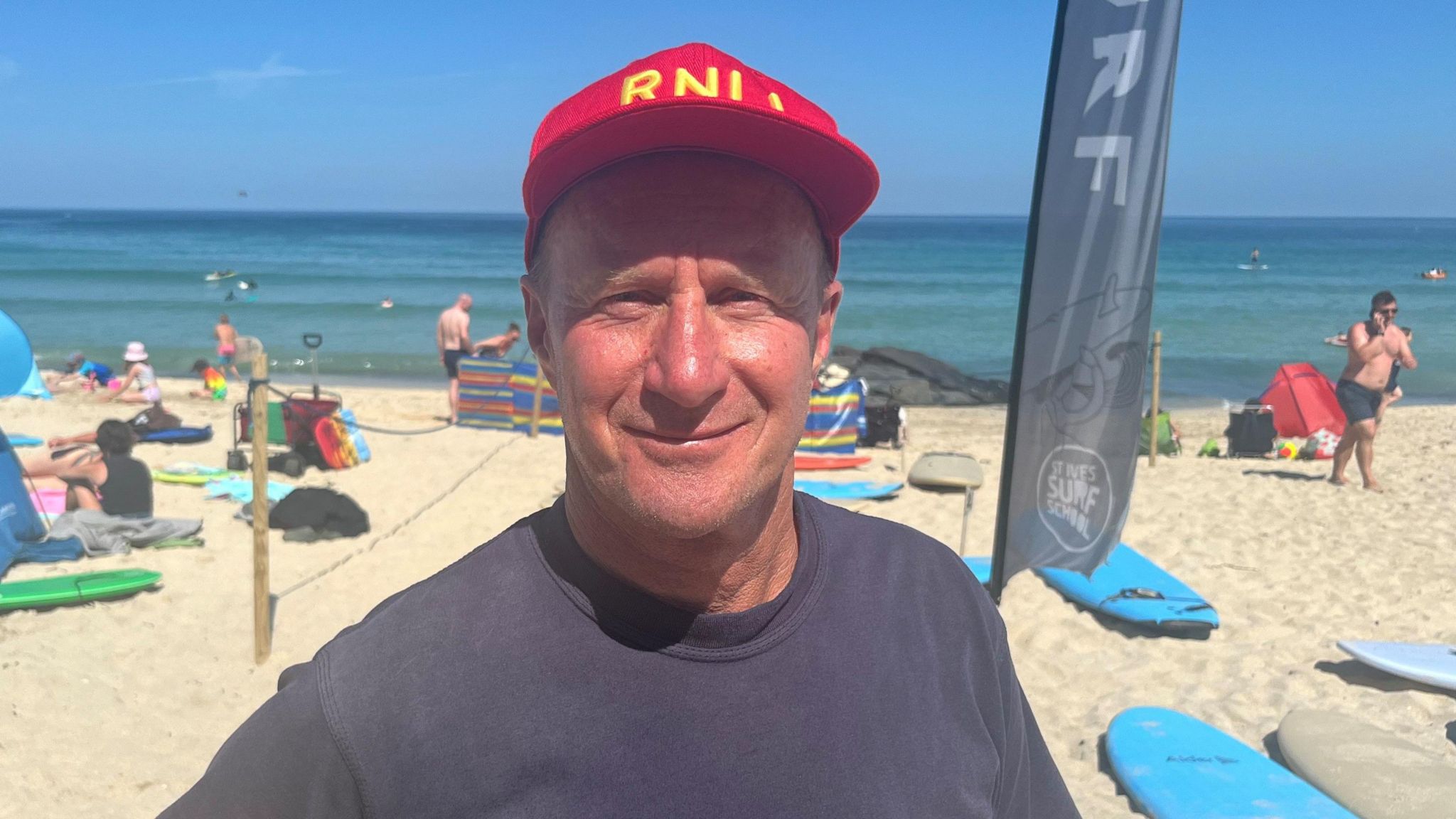 Tanned Tim Whitefield is smiling at the camera while squinting slightly at the sun wearing a dark blue T-shirt and red cap bearing the letters RNLI in yellow, the beach ocean and people are visible behind him