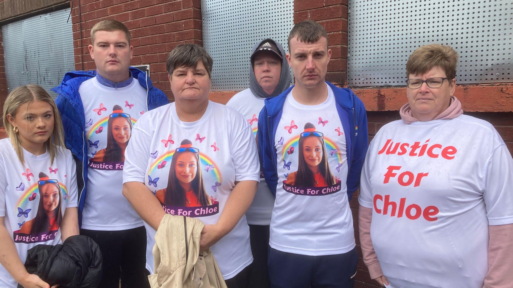 Chloe's family standing in front of the red brick boarded up windows wearing white t-shirts which have a picture of Chloe who has brown hair and a rainbow and butterflies in the background. The t-shirts say 'Justice for Chloe'. 

Left to right: 
Blond middle parting to shoulders young woman with earrings, Blond short hair young man with blue hoody over t-shirt, Middle aged brown short haired woman holding cream jacket, Middle aged man wearing grey hoody under t-shirt and a cap, young man with short brown hair wearing blue jacket open and middle aged woman with short brown hair and glasses with a different t-shirt to the rest which is white with capital letters in red 'JUSTICE FOR CHLOE' written on it.  