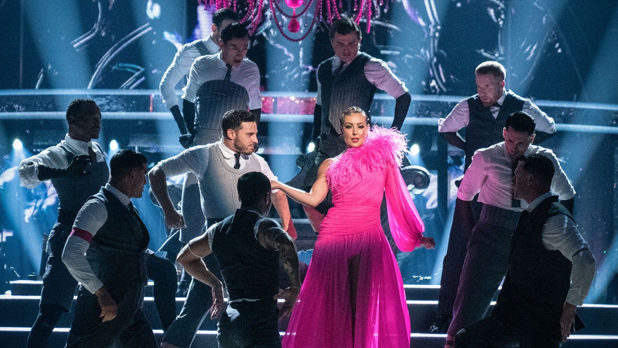 Amy Dowden wears a pink dress with feathers around the neck line as she dances down steps in the Strictly Come Dancing studio surrounded by the male professional dancers