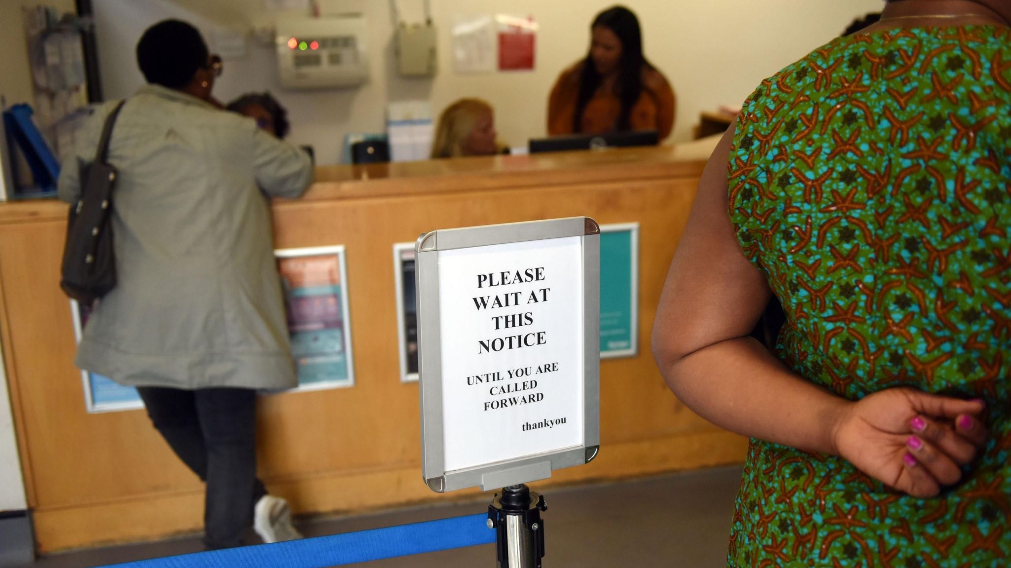 A sign at a GP's reception desk, that says please wait at this notice until you are called. Next to the sign is a person standing at the desk speaking to receptionists. 