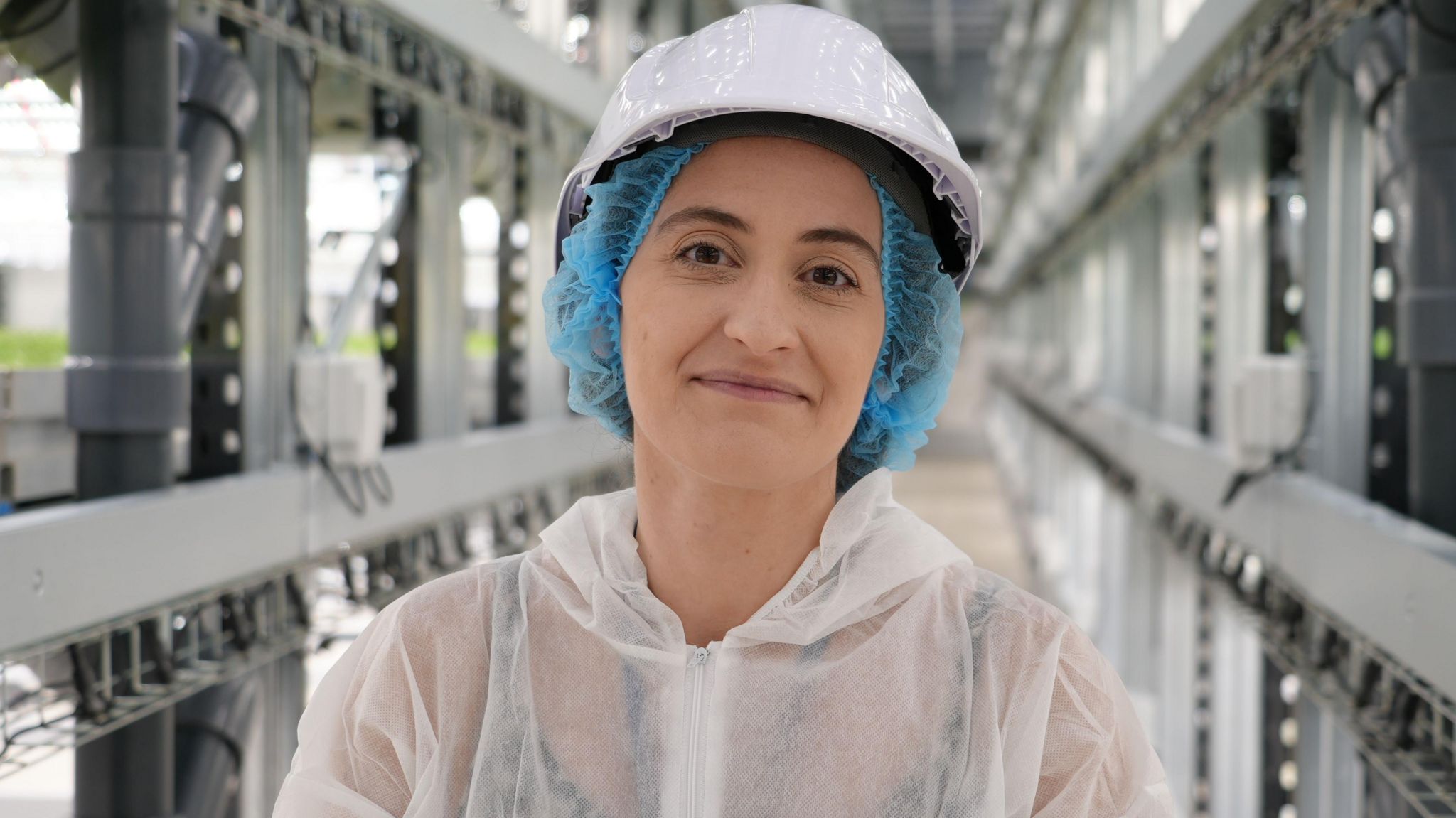 A woman wearing a white plastic suit, blue hair net under a white helmet inside a factory