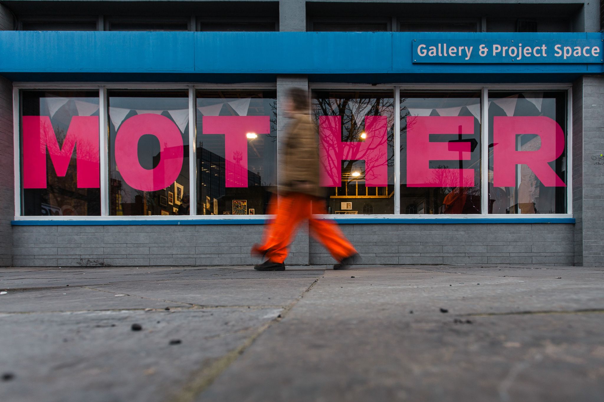 blurred figure walking past a sign saying "mother" in the windows of an exhibition space