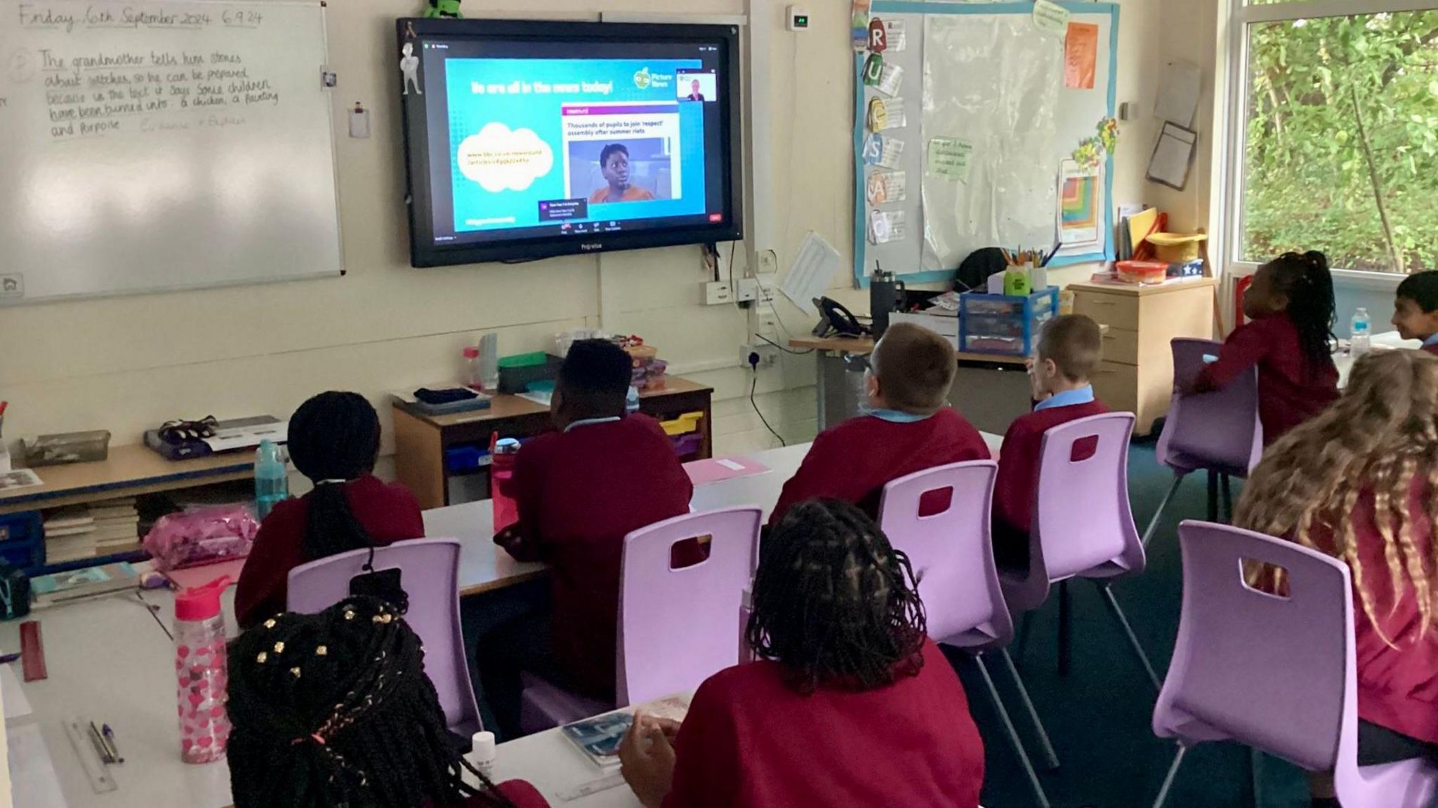 Primary-aged children in red jumpers  sit at rows of desks with their backs to the camera. The Big Back to School Assembly is being shown on the interactive whiteboard on the wall.