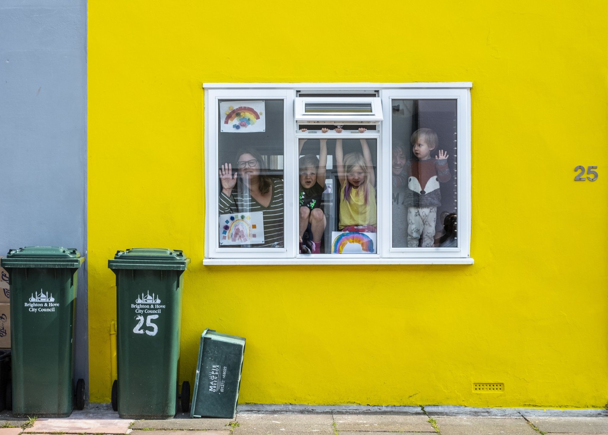 A woman and three children playfully look through their window - which has pictures of rainbows in it. Their window is surrounded by a bright yellow wall with the number 25 on it. The adjacent property is blue. Out front on the pavement there are two green wheelie bins and one smaller green recycling box lying on its side. 