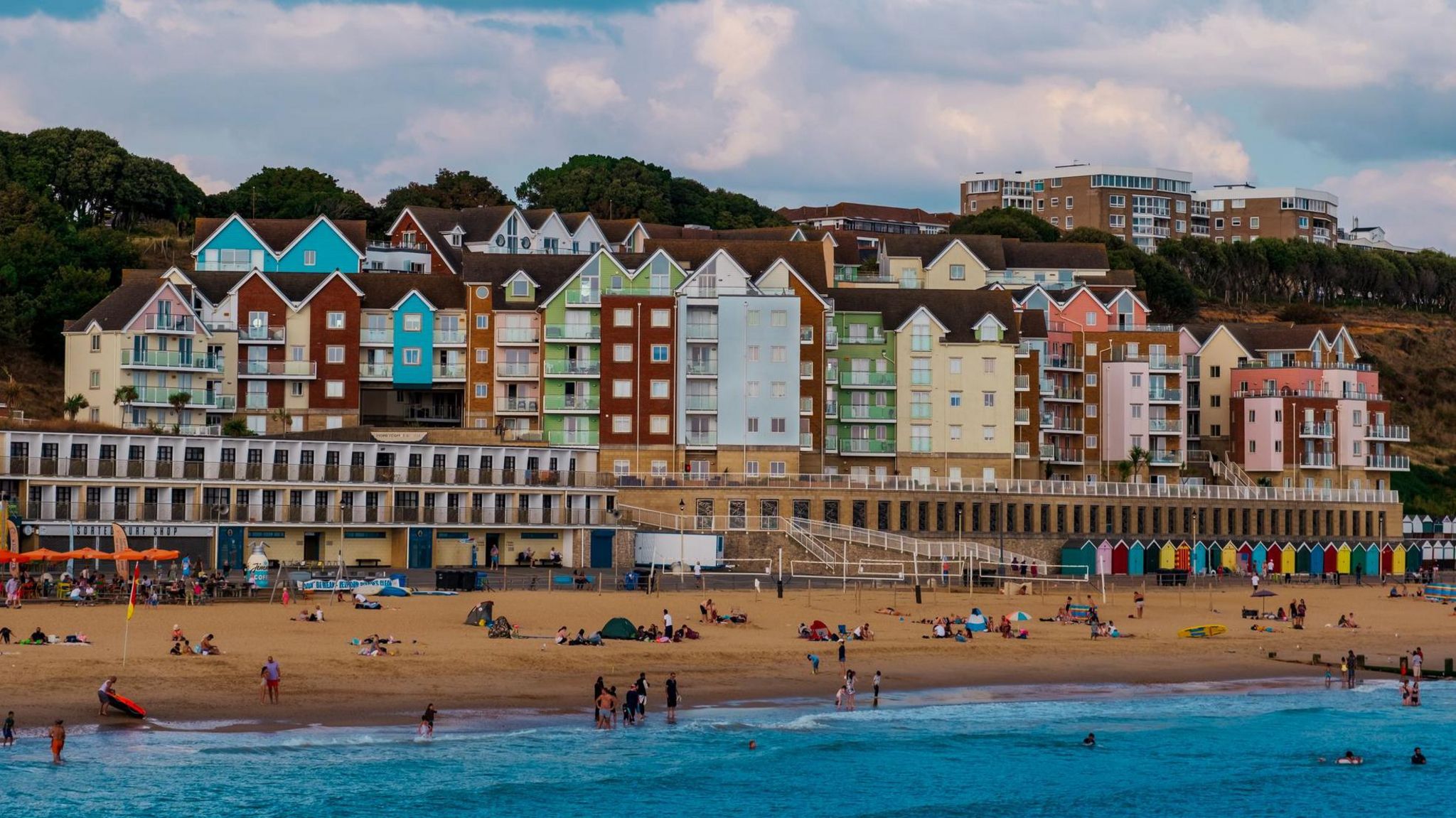 Dozens of people lay on a sandy beach. There are colourful tall houses beyond, as well as beach huts. There are some people in the sea.