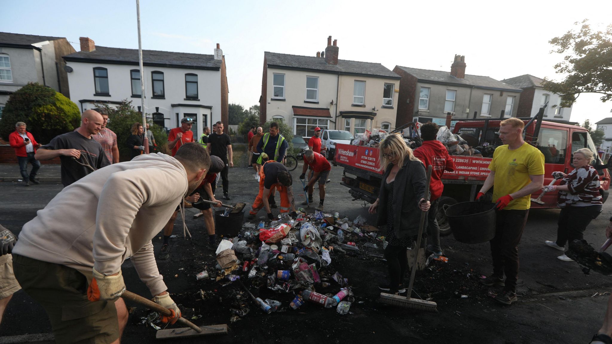 A number of people with brushes make a pile of debris including cans and bottles.