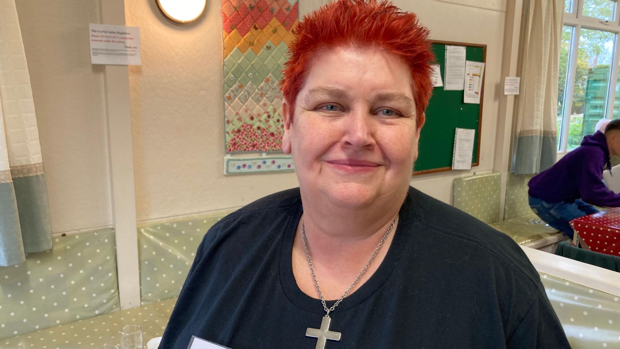 Volunteer Heather Windows with very short red hair wearing a cross and a dark top in a hall smiling at the camera