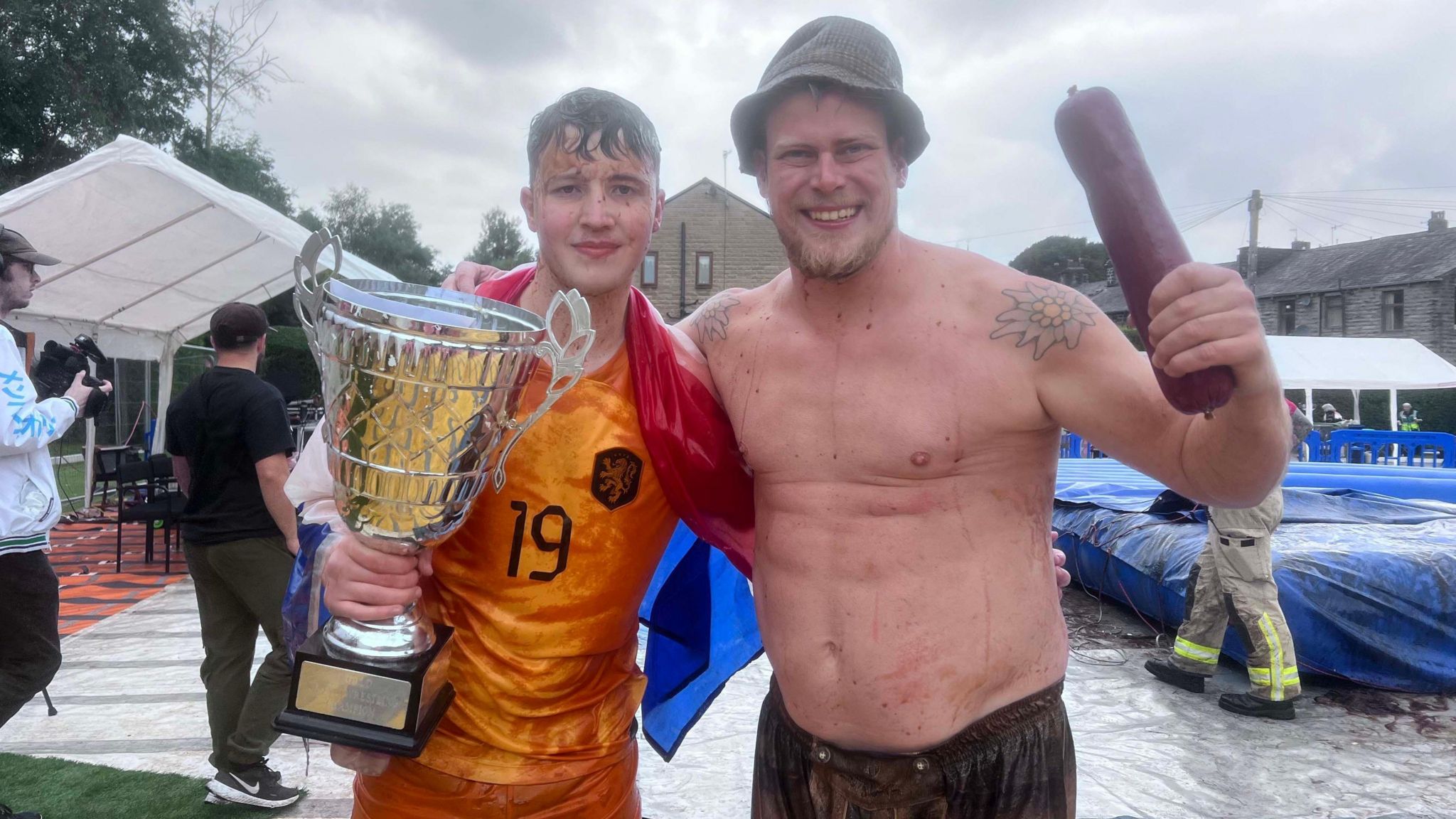 George Young (left), the teenage winner of the World Gravy Wrestling Championships, with a fellow competitor, at the Rose 'N, Bowl, a pub in Stacksteads, Rossendale. George is holding a trophy with a gravy-soaked face next to a man who is topless holding a sausage bat wearing a bucket hat. 