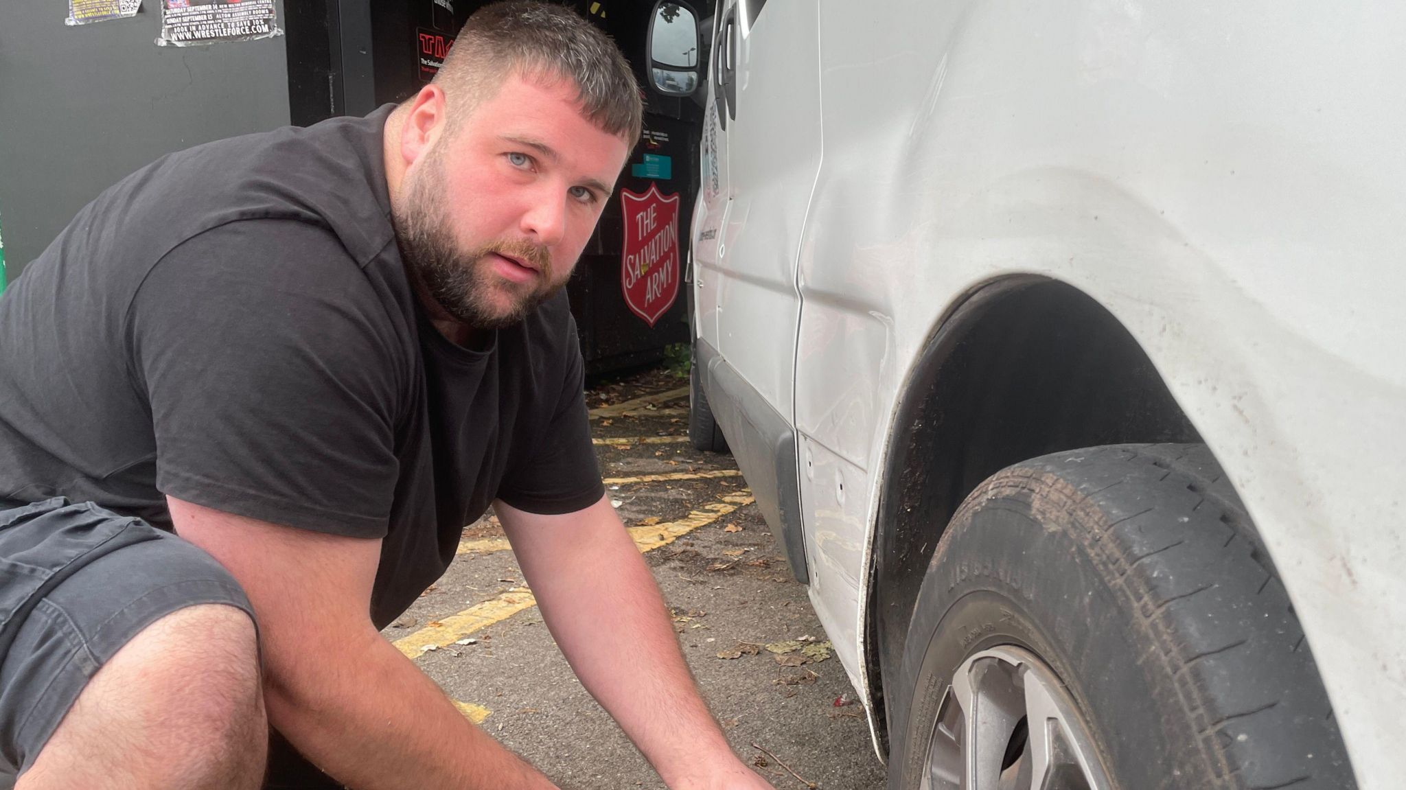 A man is crouched next to the tyre of his white work van. He is looking towards the camera and is wearing black shorts and a black t-shirt. He is red in the face. 