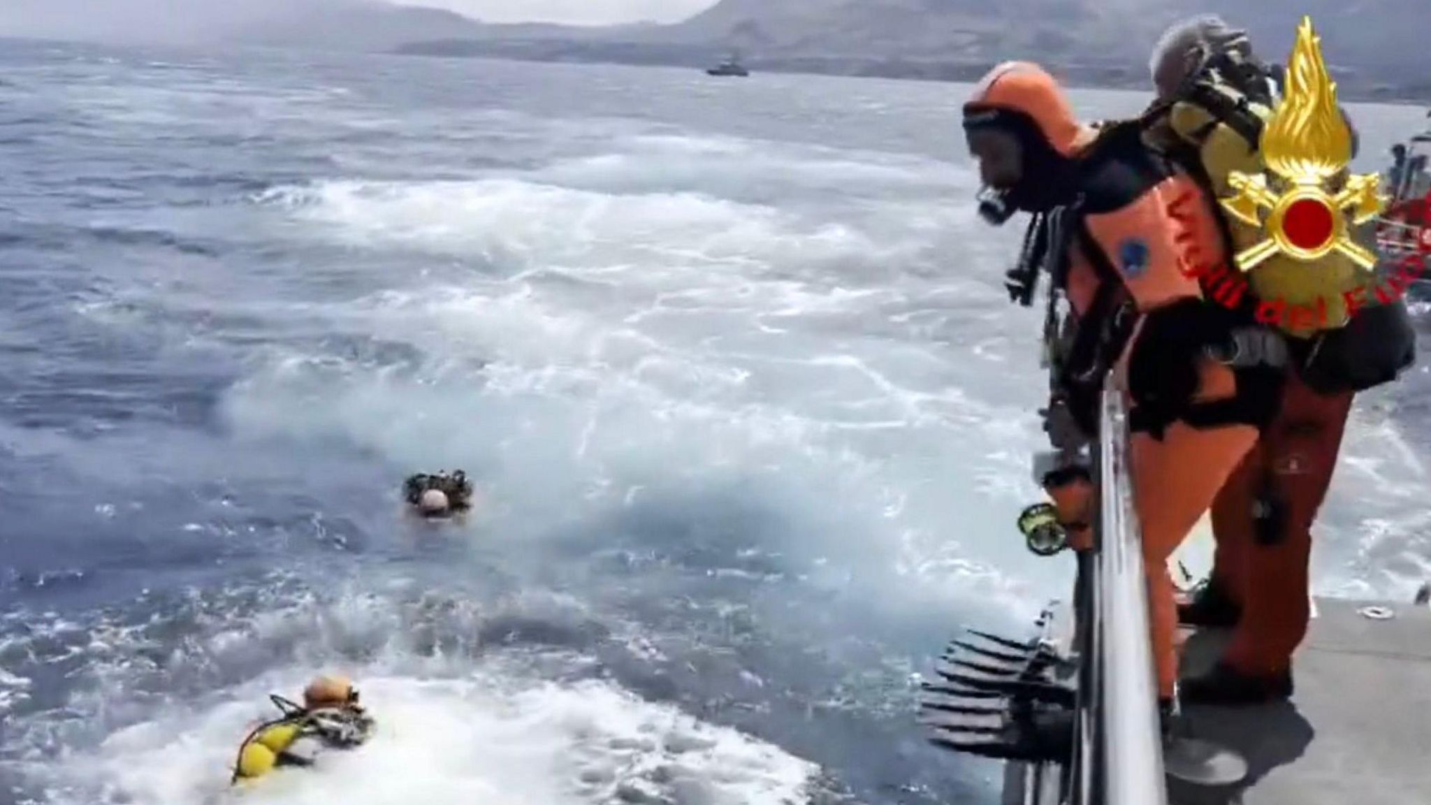 Diver pictured in water with waves around him as another diver - dressed in orange diving suit and flippers with an oxygen tank and mask on - prepares to jump into the water