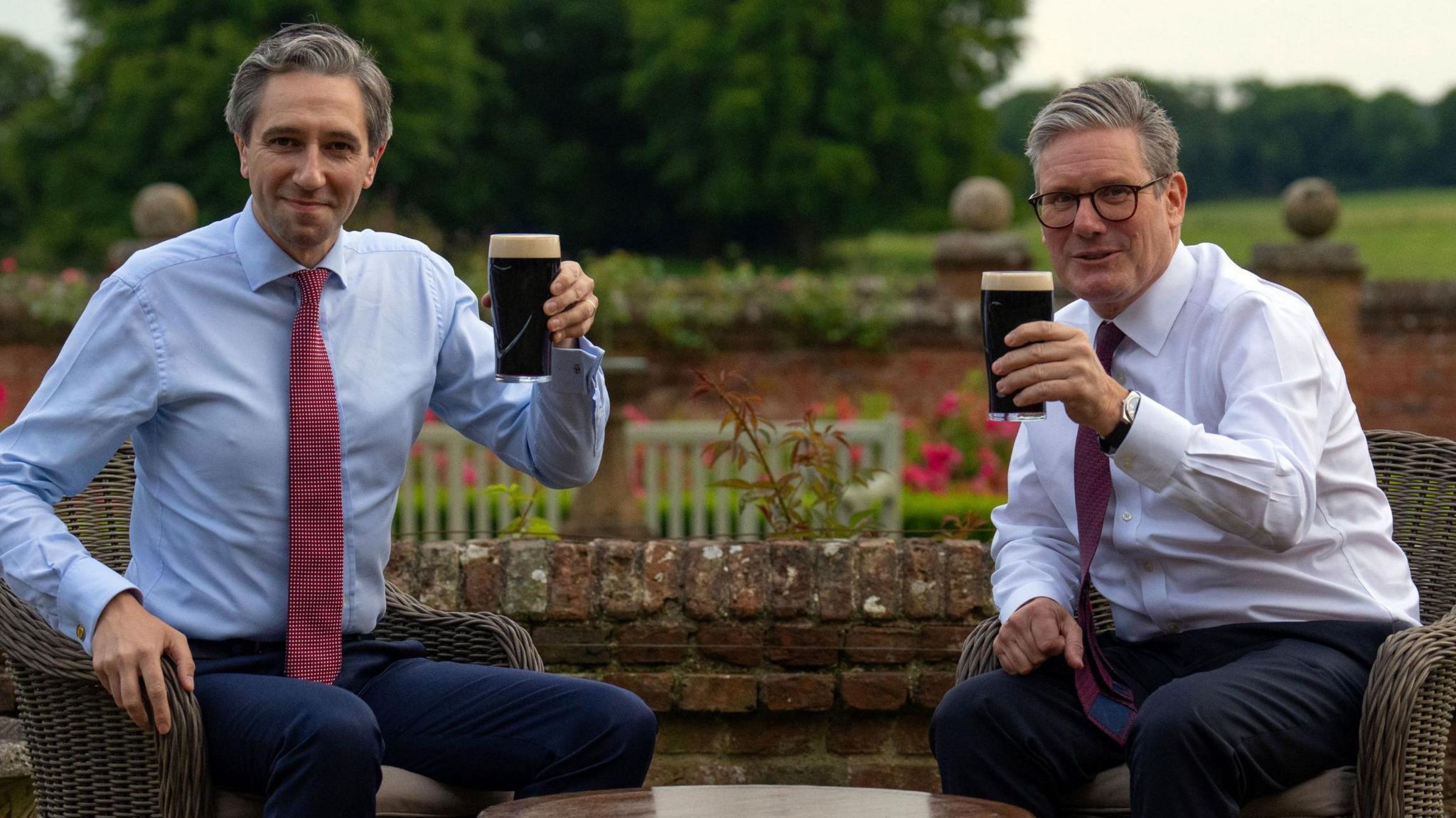 Simon Harris and Sir Keir Starmer in shirts and ties raising full pint glasses of Guinness in the direction of the camera. They are sitting in wicker chairs with roses and trees in the background, and are on either side of a round wooden table