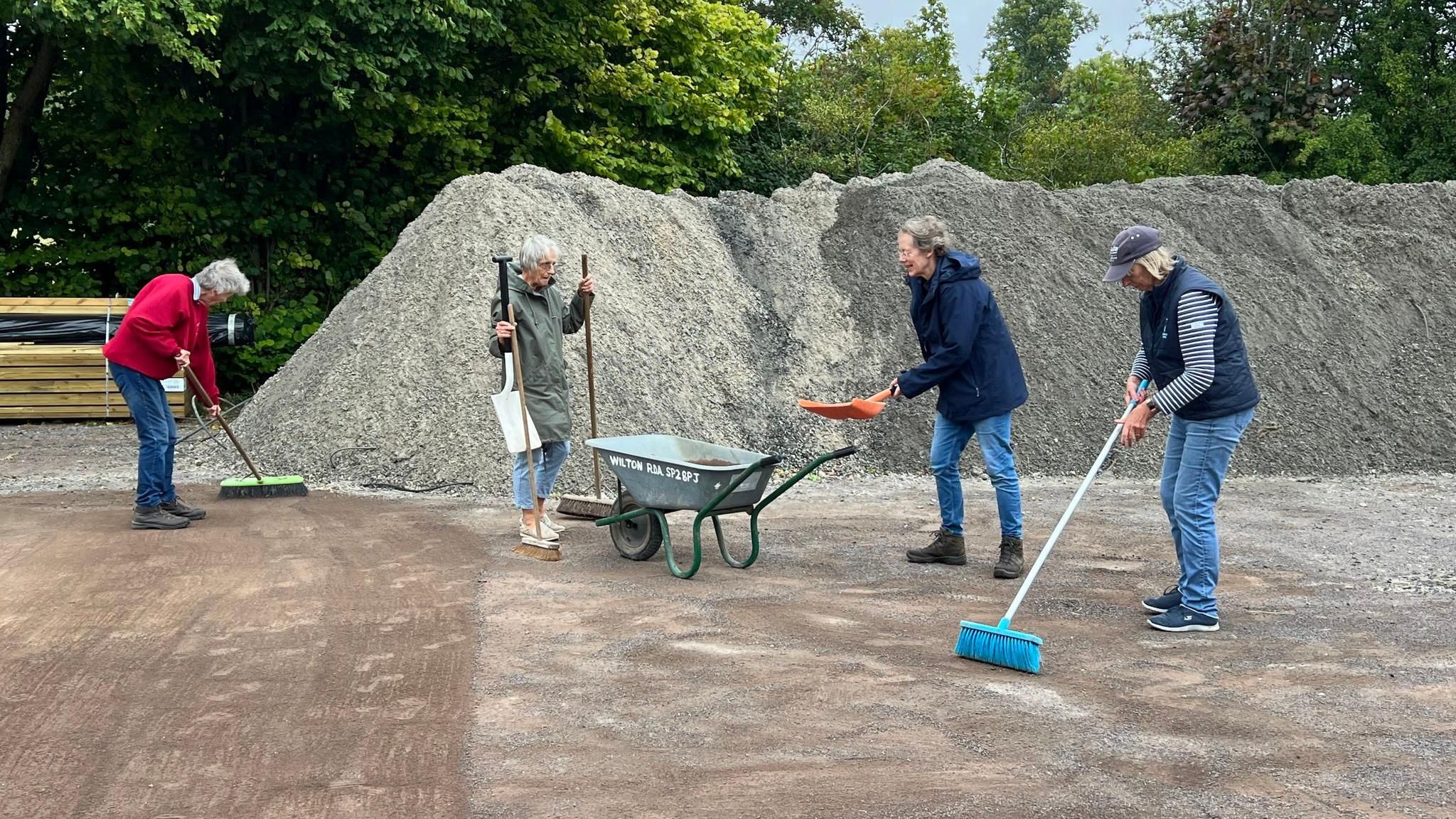 Group of people outside with brooms and a wheelbarrow with a large pile of gravel behind them