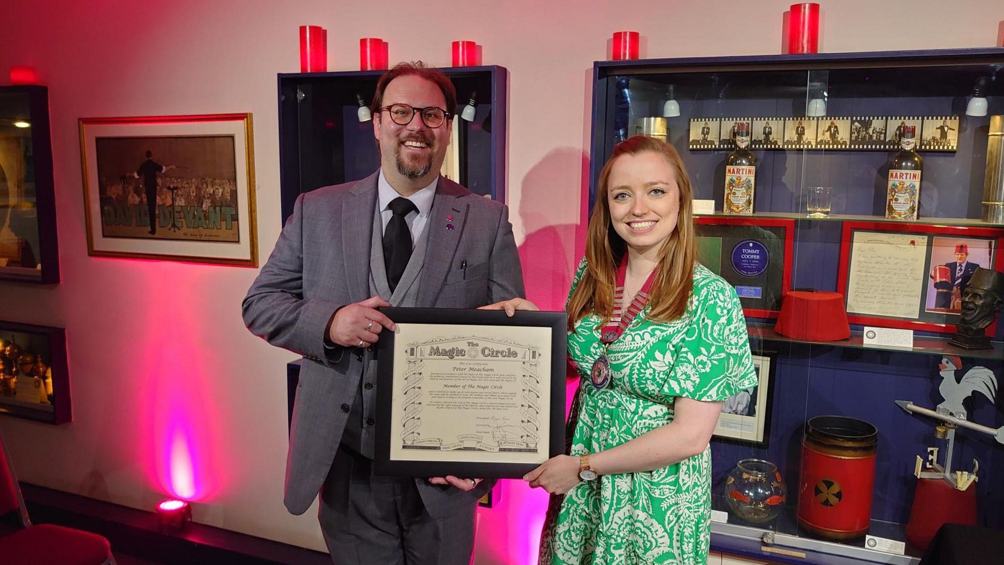 Peter Meacham standing with a female friend, the two of them holding a framed Magic Circle certificate, with a display cabinet behind them