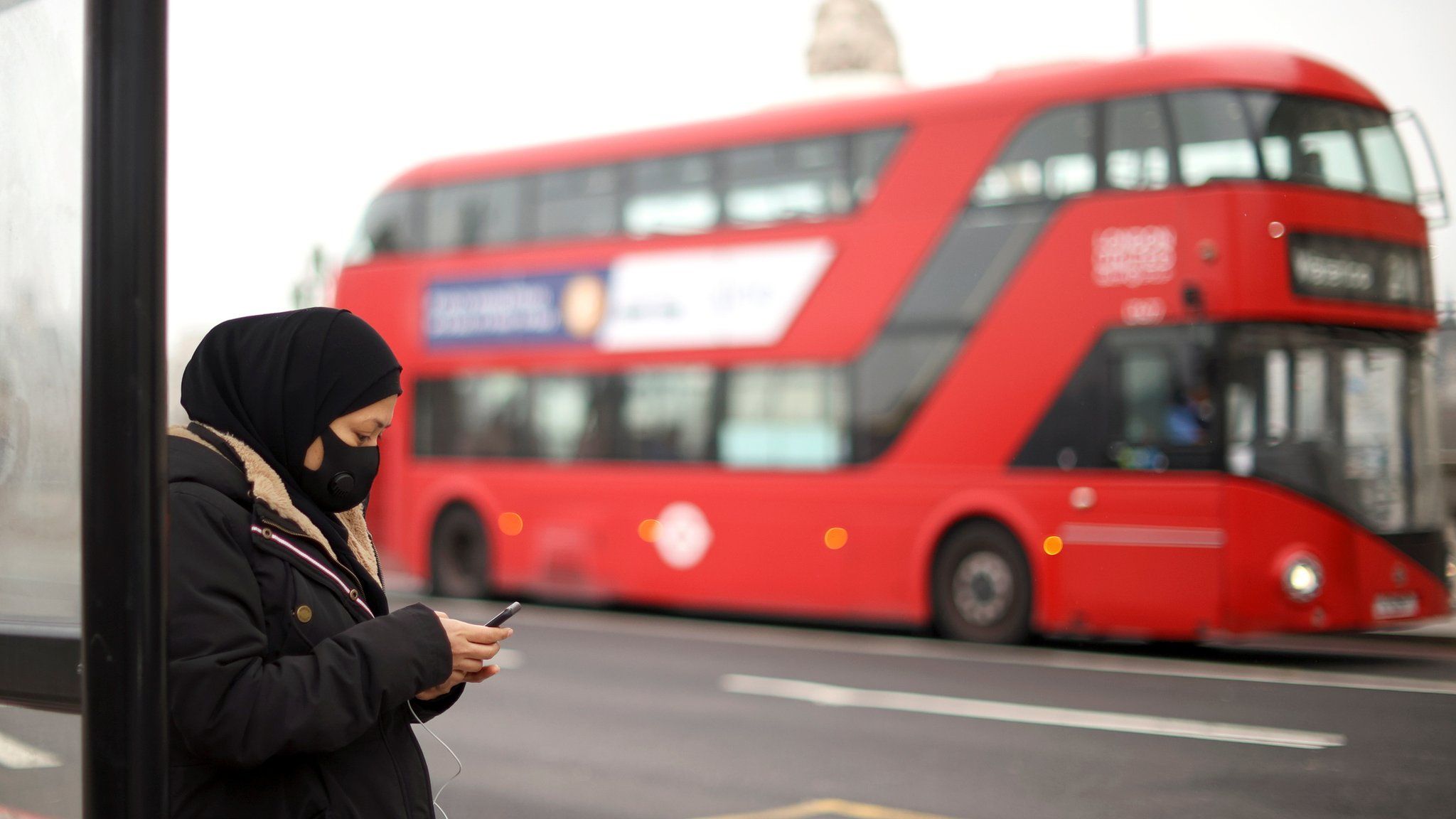 A woman wearing a protective mask uses her mobile at a bus stop