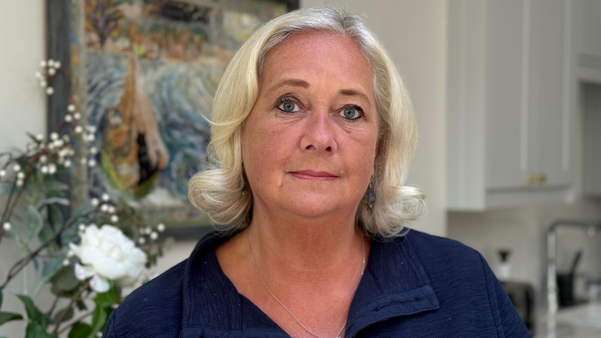 Tess Jones has grey blond hair and is wearing a blue open-necked top. Behind her are cream flowers and a multi-coloured image on the wall.