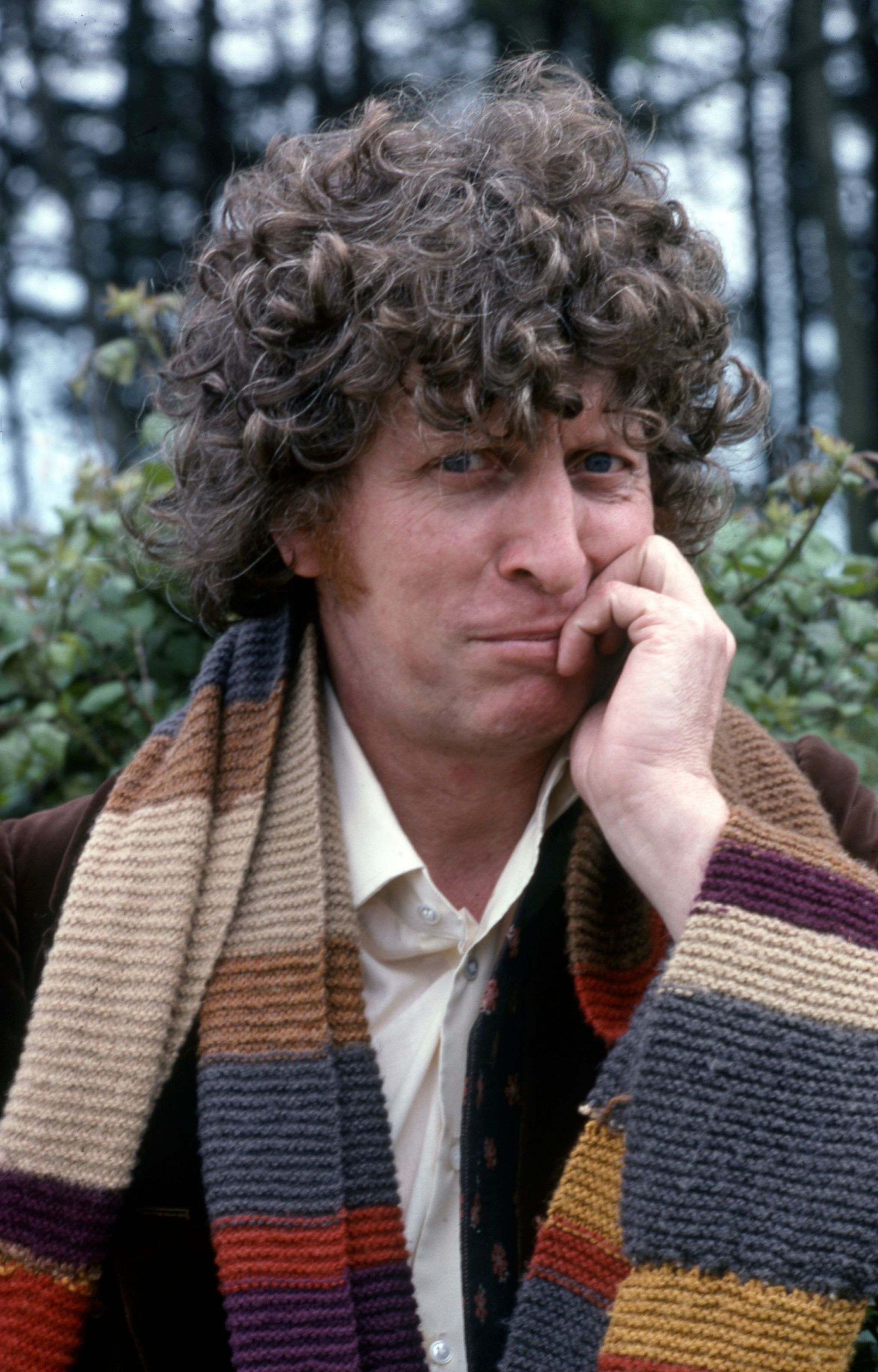 Tom Baker in costume as the 4th Doctor, looking at to camera with the beginning of a smile