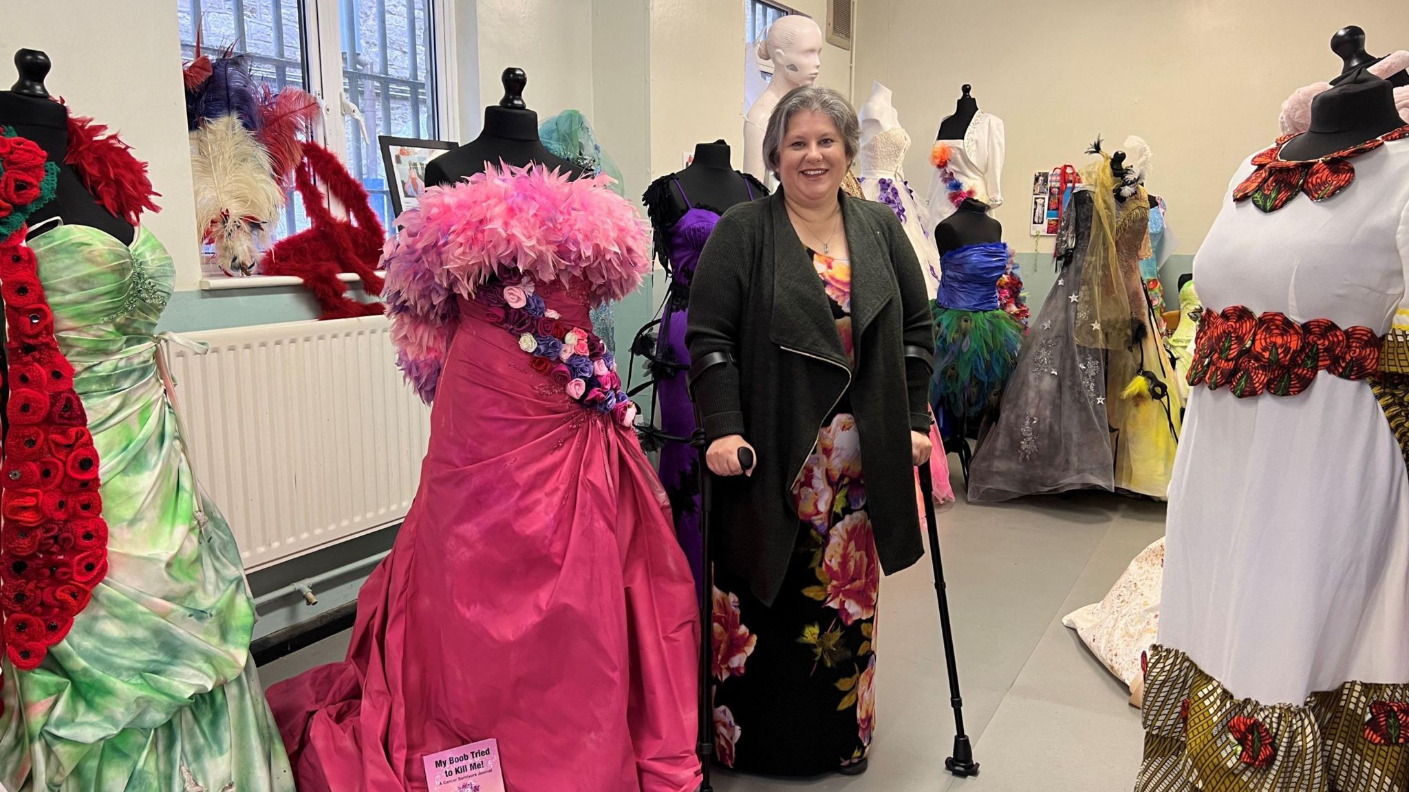 Lisa Holman standing next to the pink dress she made. The dress has pink feathers around the top and a band of colourful flowers in a diagonal line down the front of it.