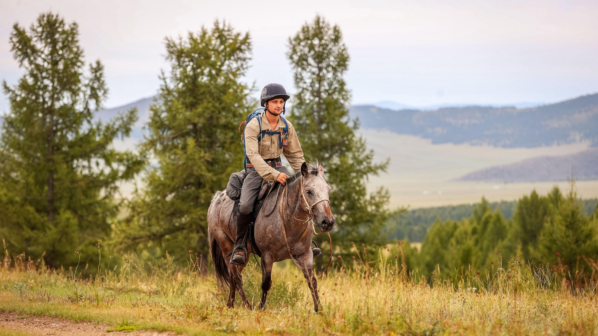 A young man on horseback. The horse is dappled brown and white, and is running. The young man, Charlie is wearing a black helmet. Under their feet is grass, and they are surrounded by green trees and bushes, with mountains and hills in the background