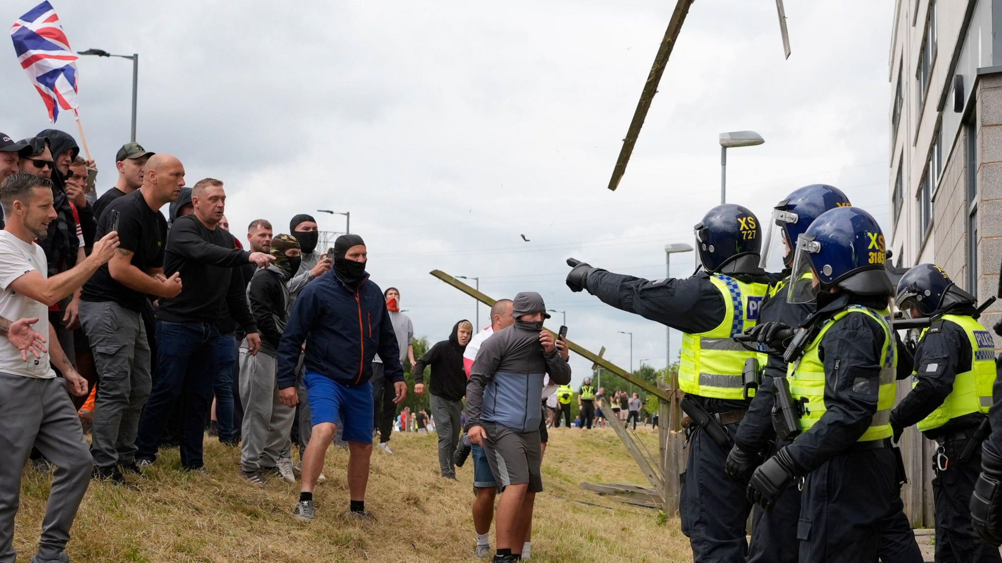 A line of protesters, some of which are wearing face coverings, facing off police officers outside the hotel. Some protesters can be seen pointing at officers and filming them with mobile phones.