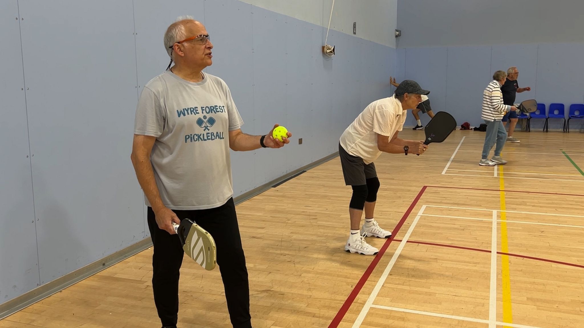 A man wearing glasses is holding a yellow pickleball and paddle. He is in a sports hall with a wooden floor. Behind him are three other pickleball players. They are holding up paddles.