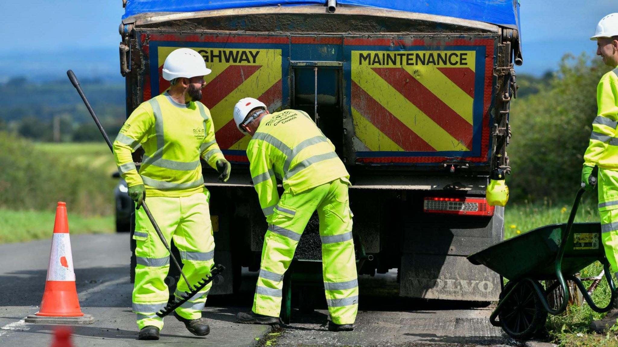 Three workman repairing the road. A lorry carrying asphalt is parked behind them.