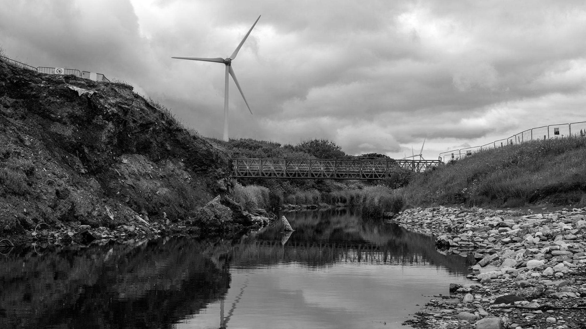 A black and white picture with a river in the foregound a bridge in the middle ground and a turbine in the background 