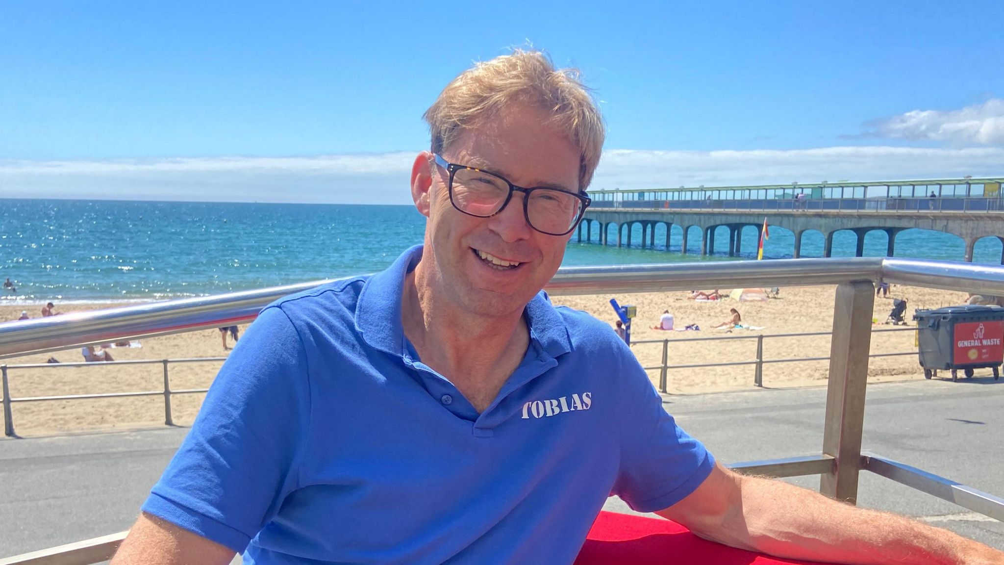 Conservative Tobias Ellwood wears a blue polos shirt with his name on it and dark-rimmed glasses